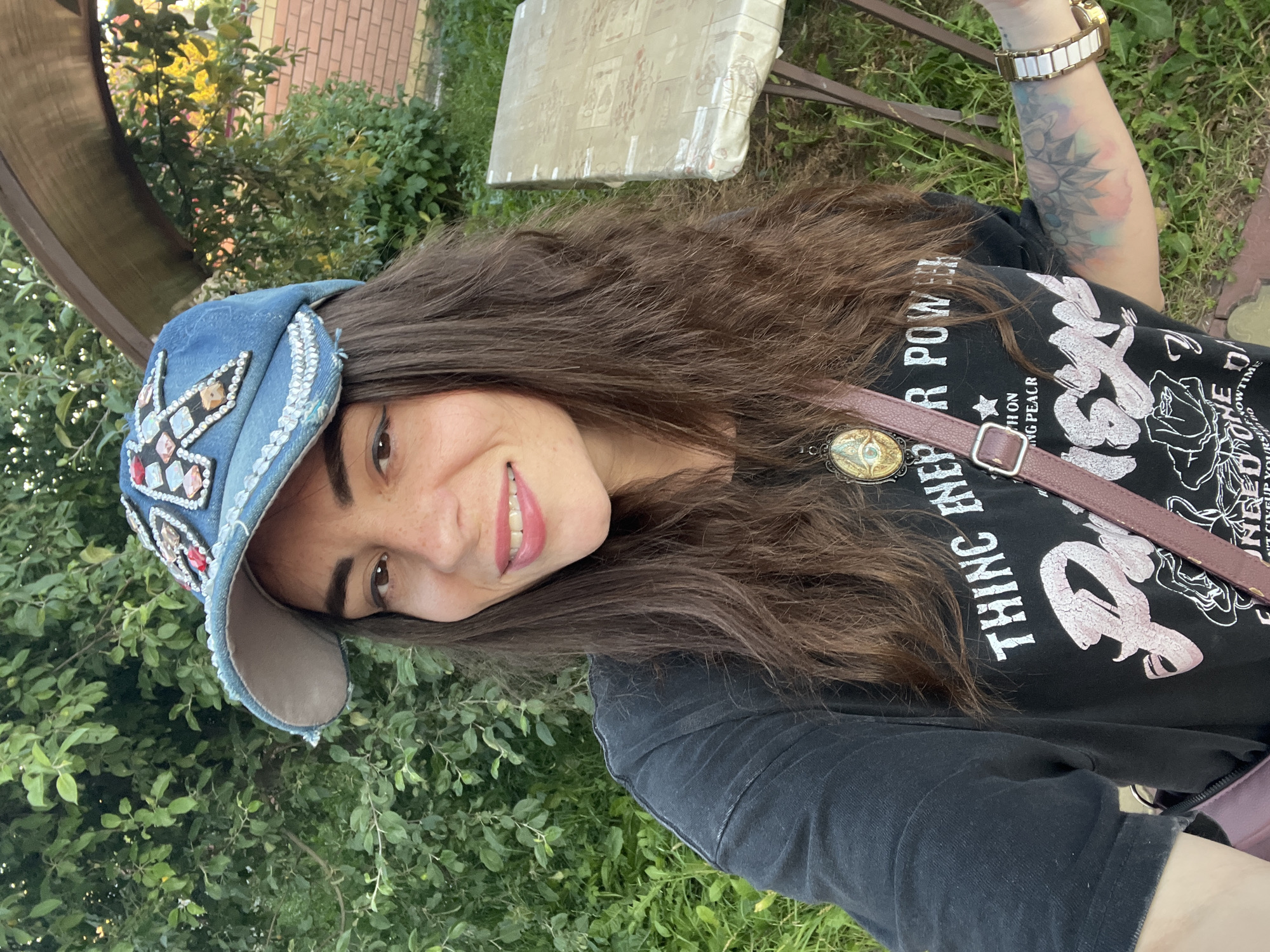 A woman is taking a selfie outdoors. She is wearing a denim baseball cap adorned with rhinestones and beaded embellishments, a black t-shirt with a graphic print, and a burgundy crossbody bag. She has long, dark, wavy hair and is wearing a watch on her left wrist. A necklace with an amulet is visible. The background shows greenery, including trees and bushes, a brick wall, and a table covered with a patterned tablecloth. The scene appears to be in a residential area or garden.