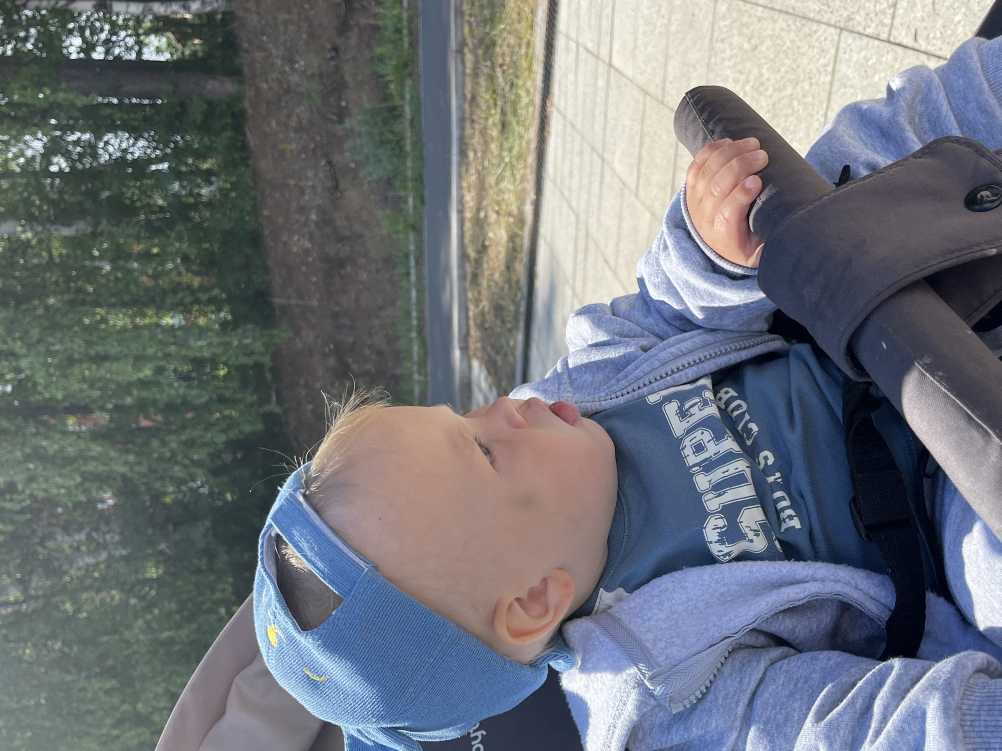 The image captures a baby in a stroller outdoors in Chaikovsky, Russia, on a bright, sunny day. The baby, with fair skin and light blonde hair, is seen from a slightly elevated perspective, looking upwards and to the left with their mouth slightly open and tongue visible. They are wearing a light blue denim-style hat with yellow embroidered accents, a blue t-shirt, and a grey zippered hoodie.
The baby's right hand is grasping the dark, padded handlebar of the stroller, which is equipped with a black strap system. The background features lush green trees and foliage on the left, with patches of bright light indicating sunlight filtering through leaves. To the right, a light-colored paved path with square tiles is visible, stretching into the distance. Beyond the path, there's a strip of grass and a darker, possibly asphalt, surface.
The strong sunlight suggests it is daytime with a clear sky, and the setting appears to be a park or a paved area alongside a natural environment. The baby seems curious and observant.
Visible text on the baby's blue t-shirt includes "BOY'S CLUB" and a partially visible word that appears to be "SUPERS," possibly part of "SUPERSPORT" or a similar phrase, printed in white, distressed font.
