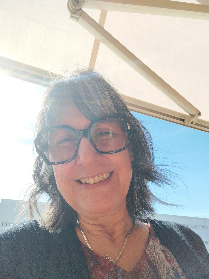 A head-and-shoulders, eye-level selfie-style photograph depicts a smiling female individual outdoors under a bright sky. The subject, appearing middle-aged or older, has shoulder-length, dark hair with prominent grey strands and wears large, dark-framed glasses. Her face displays a wide smile, revealing teeth. She is dressed in a patterned V-neck top with warm tones, covered by a dark, possibly black, cardigan. A silver-colored chain necklace with an initial-like pendant is visible around her neck. Bright sunlight from the upper left creates lens flare across the image and on her glasses. Directly overhead, a beige or white awning structure, supported by visible arms, provides partial shade. The background on the far left reveals a section of a white building with black text, partially visible as "LYCEE". To the right, a clear blue sky is visible above the upper portion of a distant, white, multi-story building with multiple windows. The scene is located in an urban area of Montélimar, France, specifically in proximity to a building identifiable as a "Lycée" (high school).