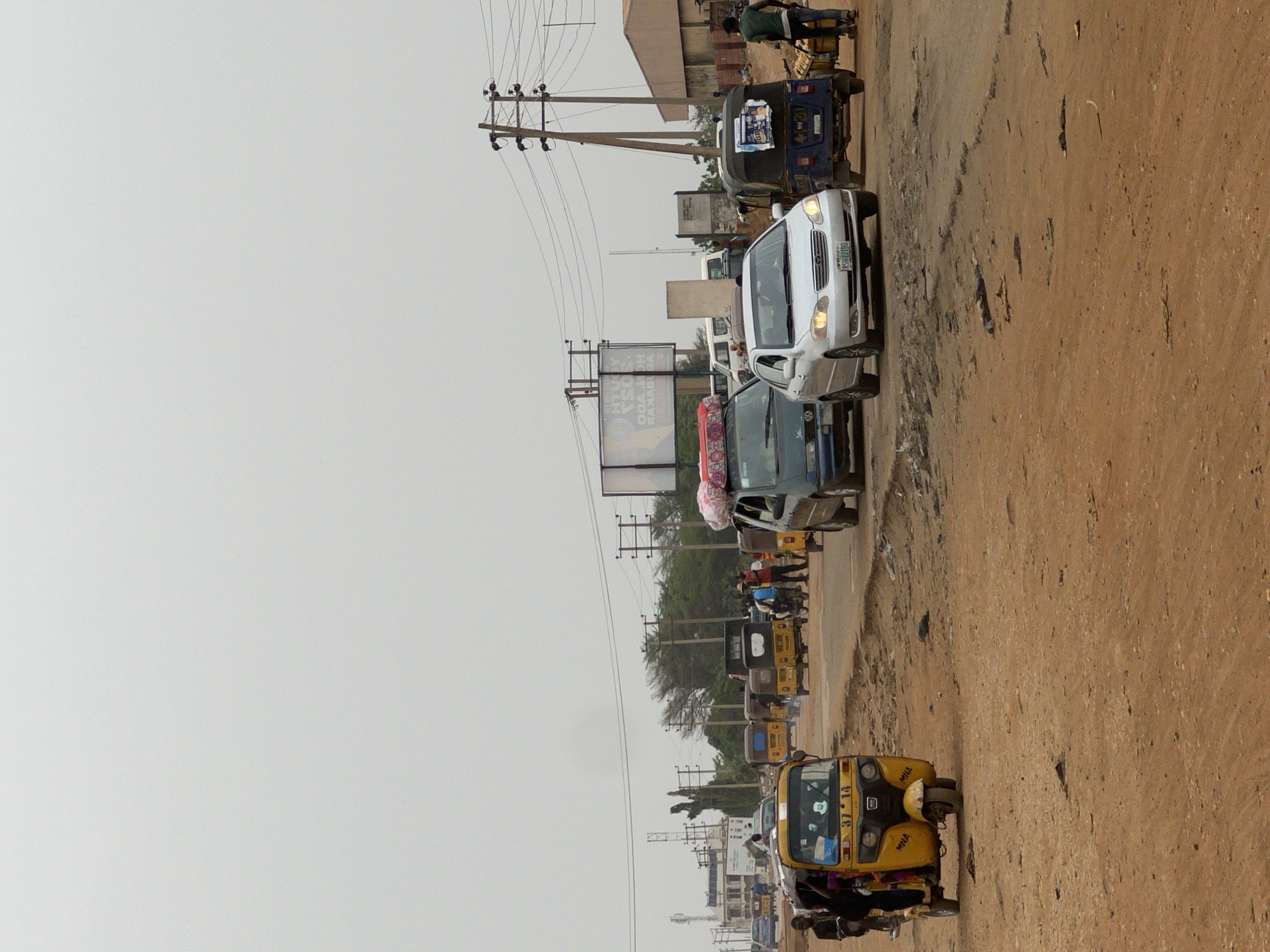 The image shows a road in Minna, Nigeria, with various vehicles, including cars and auto-rickshaws (tuk-tuks). The road appears to be in a busy area, evidenced by the number of vehicles and the presence of people. The environment consists of a mix of dirt and paved surfaces, with utility poles and overhead wires along the roadside. Billboards and other structures are visible in the background. The sky is overcast.