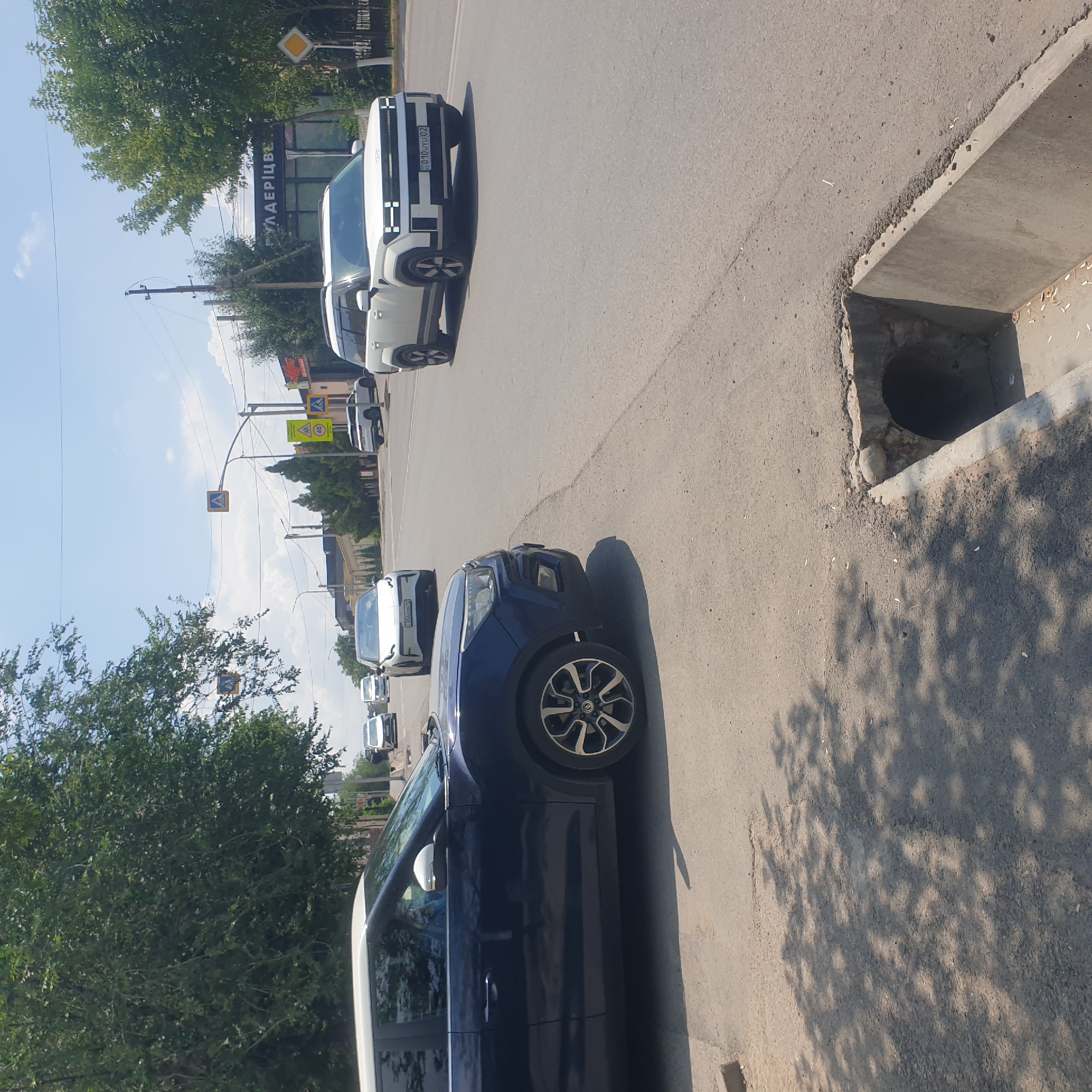 The image depicts a street scene in Shymkent, Kazakhstan, on a sunny day with scattered clouds. In the foreground, a dark blue SUV is parked on the right side of the road, partially obscuring the view of the street. Its driver's side is visible, showing a reflective blue interior. The vehicle has distinctive alloy wheels with a complex spoke pattern. To the right of the SUV, a concrete drainage ditch is visible on the edge of the asphalt road, with some debris present.
The road itself is paved and appears to be a multi-lane street. Several vehicles are driving on the road in the background. These include white SUVs, one of which has a license plate that appears to be in Cyrillic script and the numbers "010 UYU 02". There are also white sedans visible.
Traffic signs are present, including pedestrian crossing signs with yellow backgrounds and a blue pedestrian symbol, as well as a yellow diamond-shaped sign with a black border. A traffic light pole with a pedestrian signal is also visible. Buildings line the street in the background, including one with signage that includes the Cyrillic word "ГУЛАЕРЦВ". Green trees with dense foliage are abundant, providing shade on the road and sidewalk. The overall impression is of a typical urban street with active traffic and everyday infrastructure.