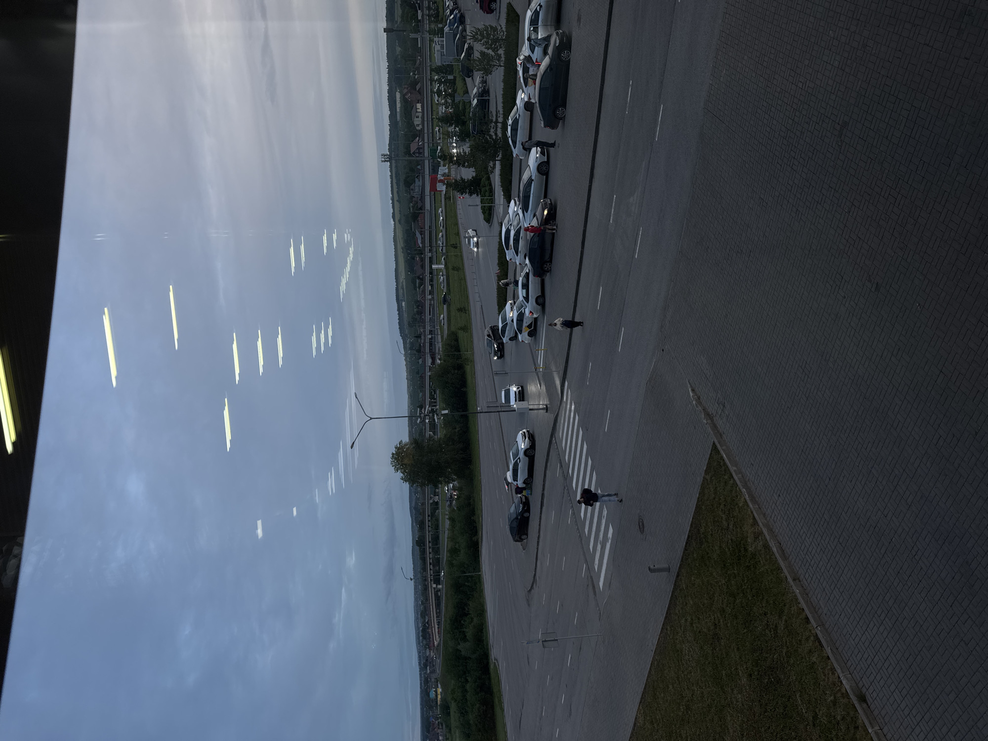 Captured from an elevated perspective, the image reveals a view of an outdoor area in Bolshoye Savino, Russia. A row of parked cars is aligned along the side of a roadway. Pedestrians are visible crossing at a designated crosswalk. In the background, structures indicative of the city are present. The sky is overcast. A section of building architecture is visible on the left side of the frame.