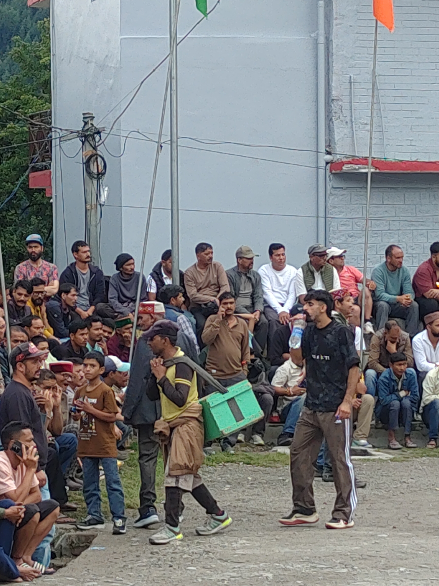 The image depicts a gathering of people in what appears to be an outdoor event in Burwa, India. The central focus is on a group of men, some seated and others standing, observing something out of frame. In the foreground, two men are walking. The man on the left is wearing a black t-shirt and brown track pants with white stripes, carrying a green container on his right side. The man on the right is partially visible, wearing a yellow t-shirt and brown shorts over black pants.
The background shows a tiered seating arrangement filled with spectators, predominantly men of various ages. Behind the seating area, a light-colored building with a grey facade is visible, along with electrical poles and wires. Two flags are discernible, one green and one orange, attached to poles. The ground is a rough, grey surface. The overall impression is that of a local event or gathering, possibly a sporting event or a public ceremony.