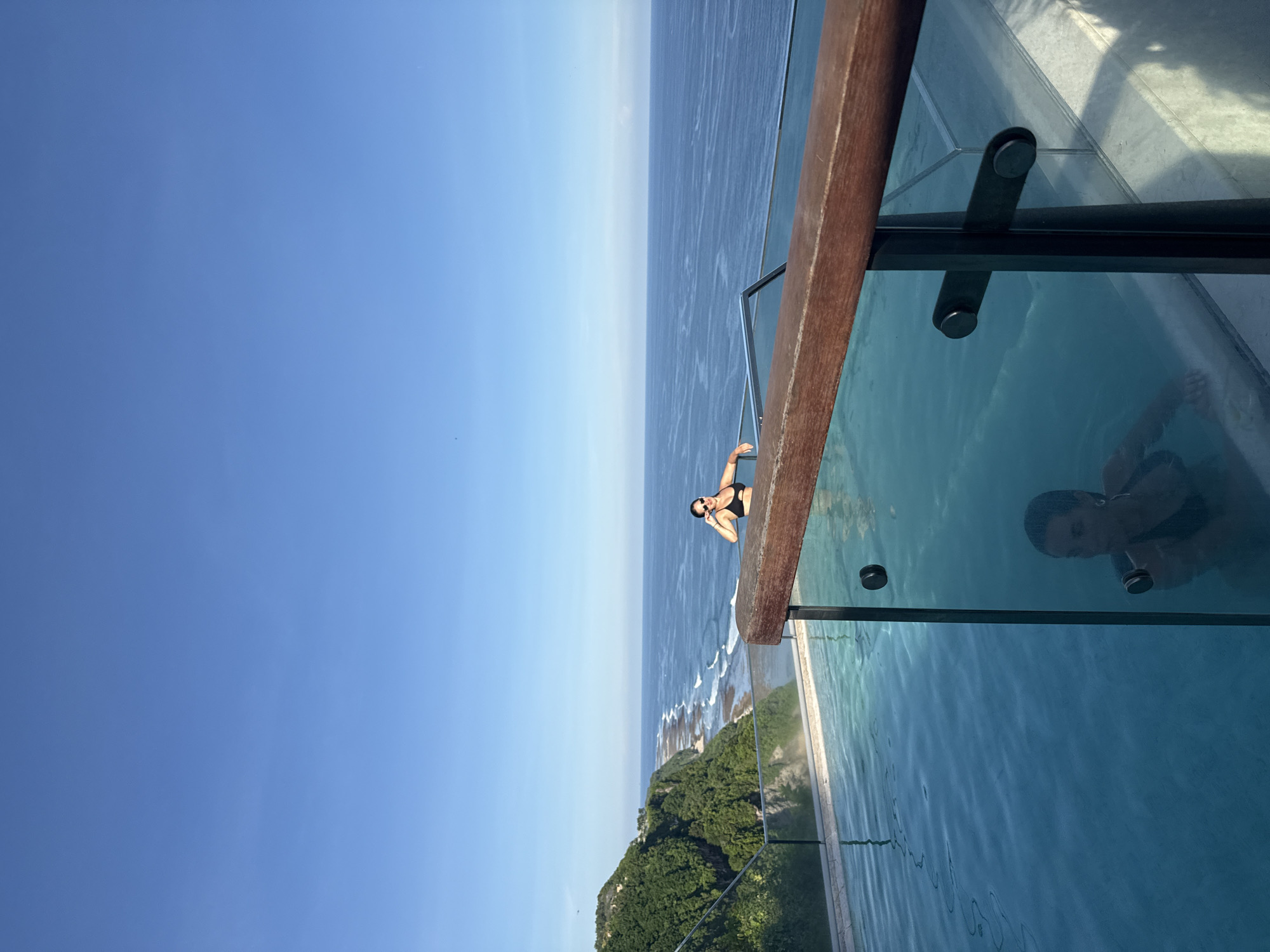 The image depicts a woman at the edge of an infinity pool overlooking the ocean in Kuta Selatan, Indonesia. She is wearing a black bikini and sunglasses, leaning on a wooden railing, seemingly posing for a photograph. The pool has a glass barrier. Her reflection is visible in the pool's water. In the background, there is a coastline with green vegetation and the blue ocean meeting a clear blue sky.