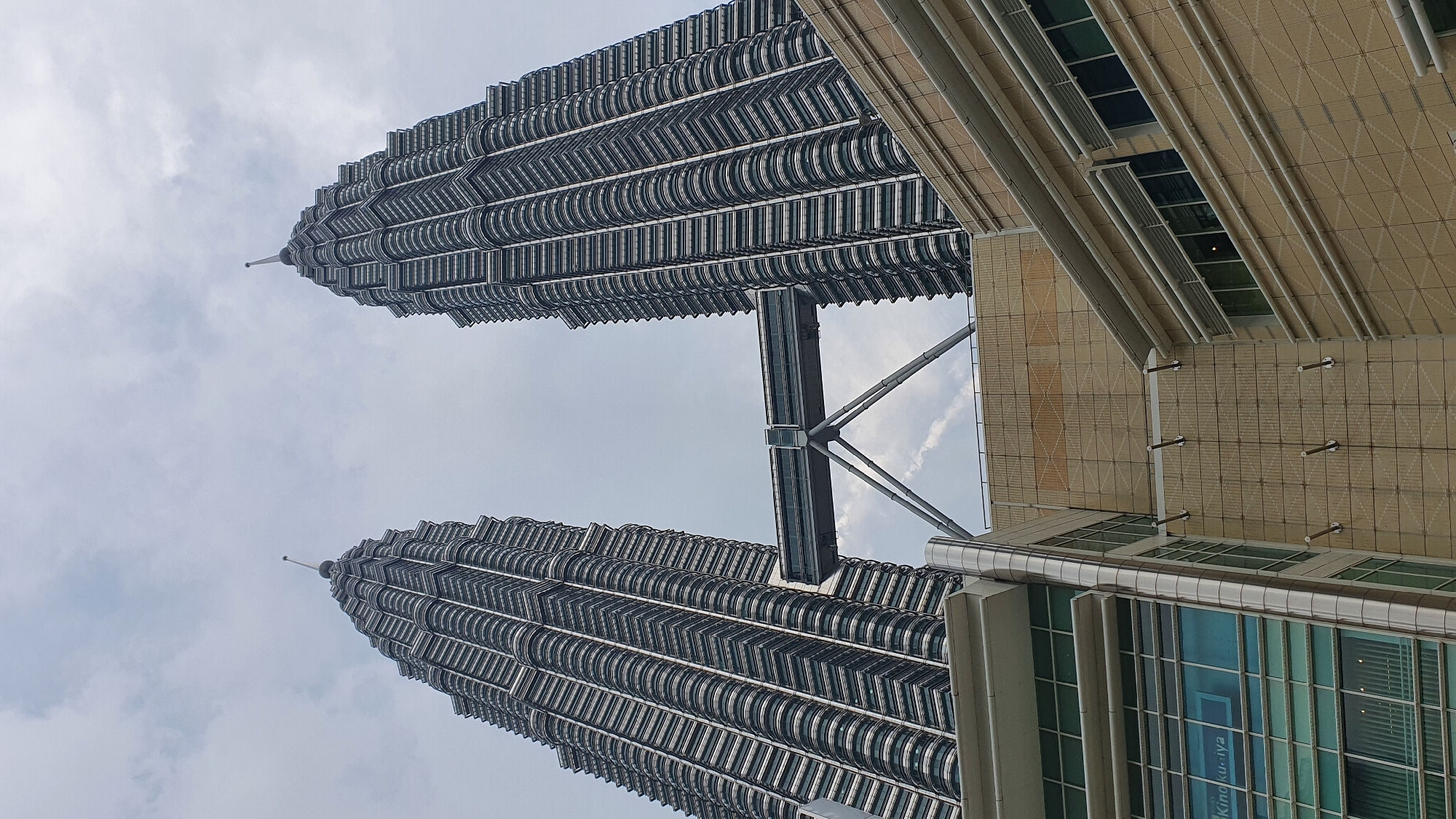 The image captures the Petronas Twin Towers in Kuala Lumpur, Malaysia, seen from a low angle looking upwards. The sky is overcast with scattered clouds. The two iconic skyscrapers, with their distinctive pointed spires and tiered facades, dominate the upper portion of the frame. A skybridge connects the two towers at a high level, supported by an intricate triangular structure. In the foreground, a portion of a modern building with large glass windows and beige tiled walls is visible. The building's facade features horizontal white piping and awnings over some of the windows. The overall scene is of urban architecture under a cloudy sky.