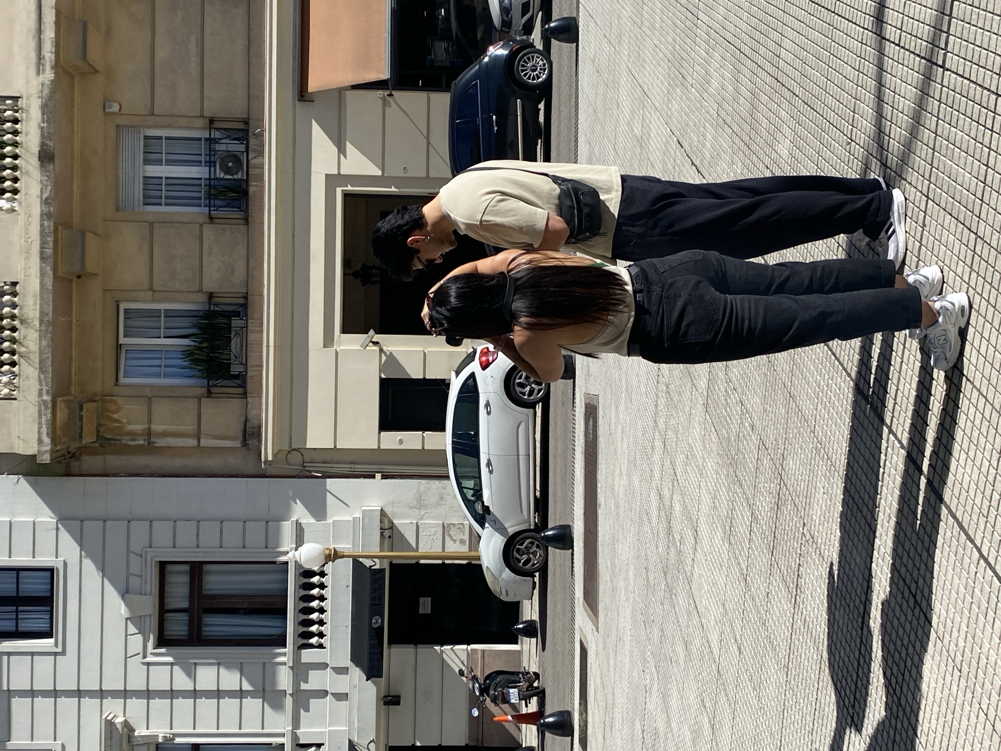 Two individuals, a man and a woman, stand on a light-colored, patterned sidewalk under bright sunlight in what appears to be a city street in Buenos Aires, Argentina. The image is rotated 90 degrees clockwise, showing the scene from an elevated perspective.
The man, positioned slightly behind the woman, wears a light beige t-shirt, dark pants, and white sneakers with black stripes. A black fanny pack is slung across his chest. He has dark hair and is looking down towards the woman.
The woman stands in front of him, facing away from the viewer. She has long dark hair and wears a light-colored sleeveless top, black jeans, and white sneakers with visible "N" branding (New Balance). Her arms are raised as if holding a camera or phone, pointing it downwards towards the ground or an object in front of her, suggesting she is taking a photograph or video.
Behind the individuals, a multi-story building with cream-colored facades, windows, and window guards is visible. A dark compact car, possibly a Fiat 500, is parked on the street behind them. Further down the street, a white compact car, possibly a Citroën C3 Picasso, is parked near a street lamp. A motorcycle or scooter is also visible on the sidewalk closer to the white car, with an orange cone nearby. Another building with white, art deco-style architectural details and dark-framed windows is on the left side of the street.
The bright sun casts prominent, elongated shadows of the people and buildings onto the tiled sidewalk, indicating it is likely midday or early afternoon on a clear day. No discernible text is clearly visible on any signs or license plates. The overall scene captures a moment of observation or photography in a bustling urban environment.