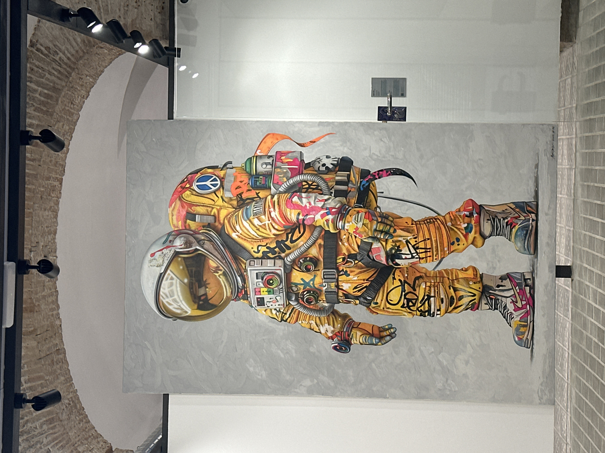 The image shows a large painting of an astronaut in a yellow spacesuit covered in graffiti. The astronaut is standing in front of a grey textured background. The spacesuit is adorned with colorful tags and images, including a peace sign and what appears to be a camera. The helmet has a golden visor that reflects the surrounding environment. The astronaut's boots are also heavily decorated. The painting is displayed in what appears to be an art gallery or exhibition space. The ceiling features exposed brick arches and track lighting. In the background, a glass partition with a door and information placards is visible. The floor is paved with grey tiles. The context suggests this artwork is part of an exhibition, likely in an urban setting given the graffiti style. Based on the architectural style and the typical placement of such art, it is plausible this scene is located in Barcelona, Spain, possibly in the El Born district, known for its art galleries and historic buildings.