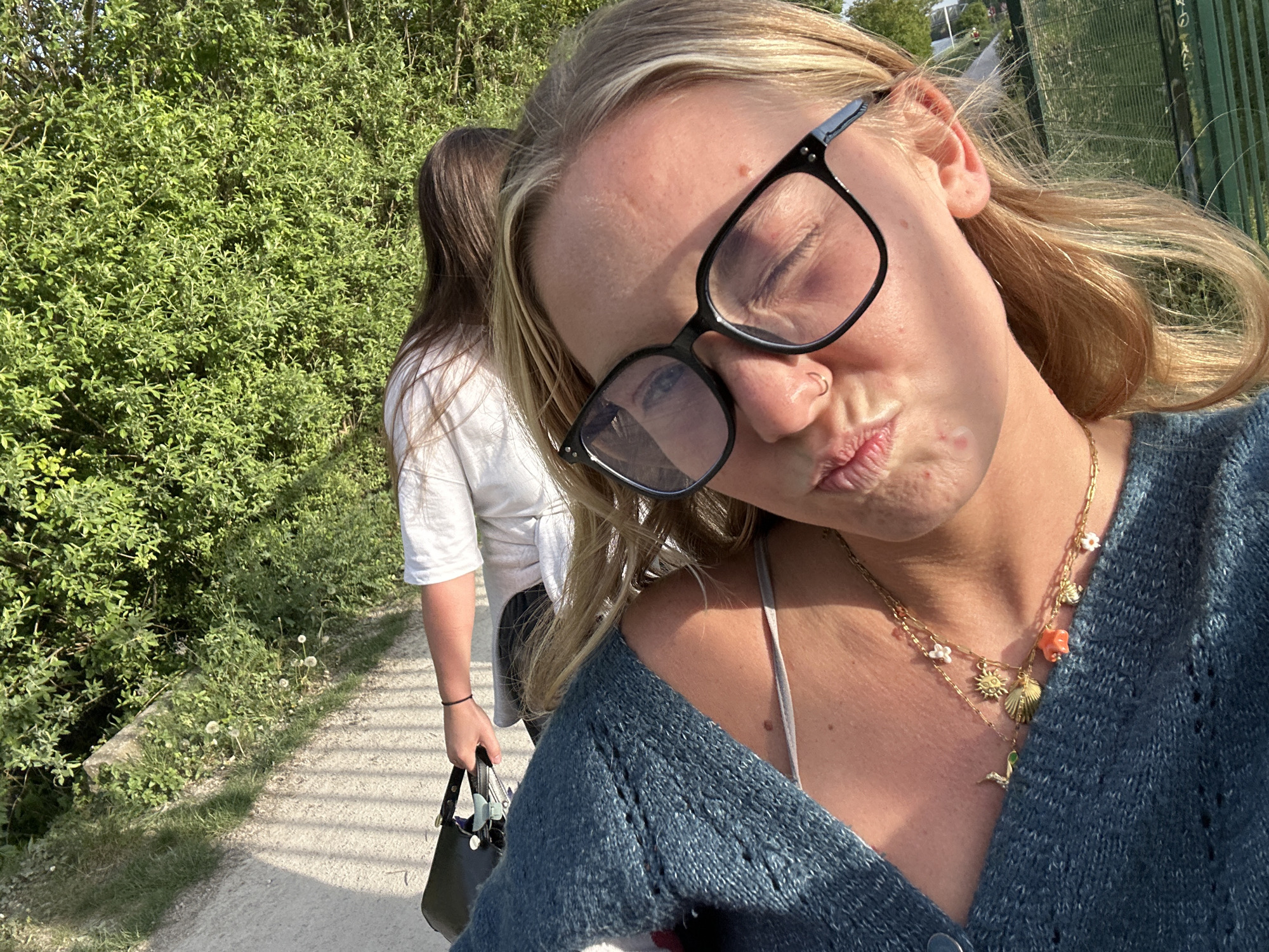 A selfie-style photograph features a blonde woman with closed eyes, wearing black-framed glasses and puckering her lips. She is wearing a blue cardigan, and a layered gold necklace with various charms. In the background, another person with brown hair and a white shirt carries a black bag. The photo appears to be taken outdoors on a paved path with green foliage on the left and a fence on the right. The location is Marquette-lez-Lille, France.
