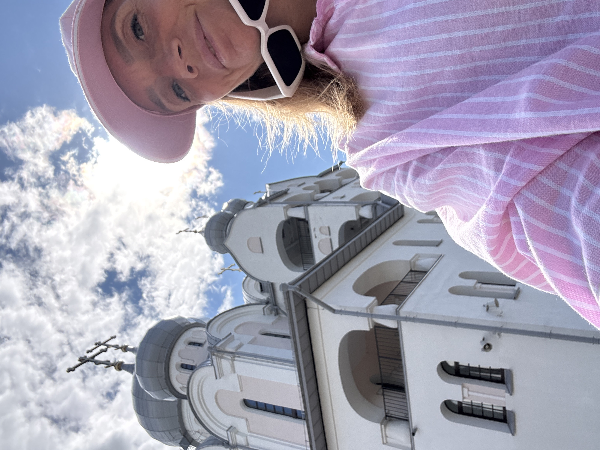 A woman wearing a pink striped shirt, a pink hat, and white sunglasses is taking a selfie outdoors. Behind her, the white facade of a large church with gray domes and crosses is visible. The sky is blue with scattered clouds. The architecture of the church suggests it is likely in Saint Petersburg, Russia, given its onion domes and cross design, common in Russian Orthodox churches. The sunlight illuminates the scene, creating bright highlights on the church and the woman's hair. The woman's hair is blowing in the wind, indicating an outdoor setting with some breeze. The overall mood is casual and sunny.