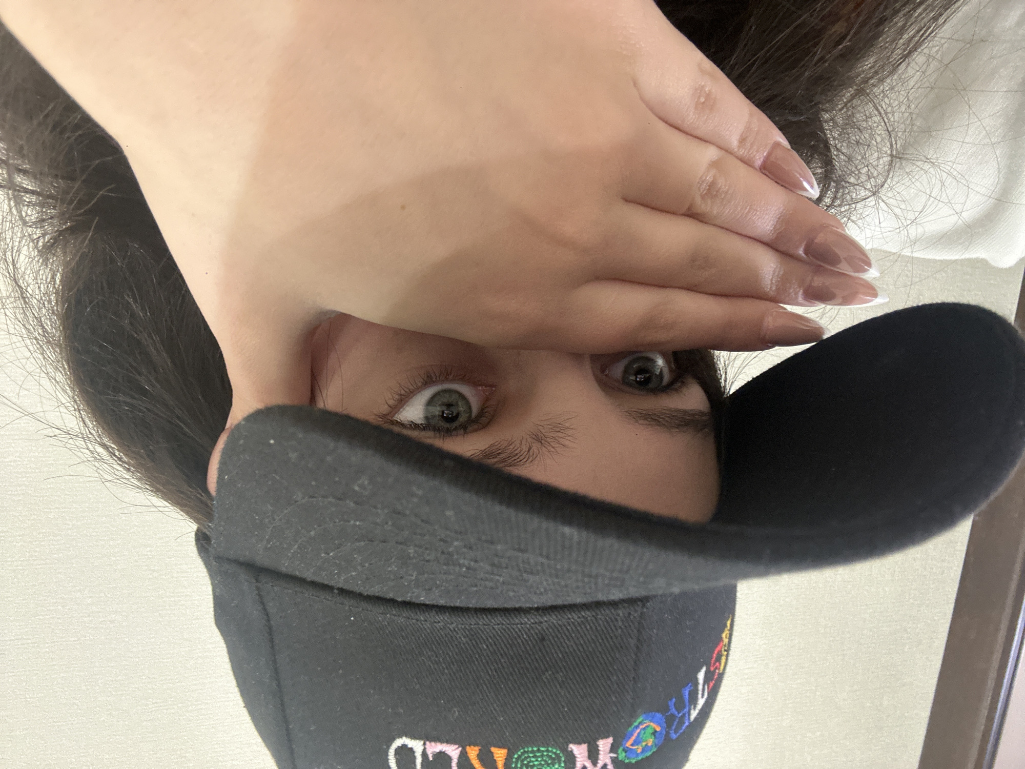 The image shows a person partially obscured by a black cap and a hand over their forehead, with only their eyes visible. The cap has colorful embroidered text "ASTROWORLD" on it. The background is blurry and indistinct, but appears to be a white wall. There is no information in the image to indicate that this was taken in Yerevan, Armenia.