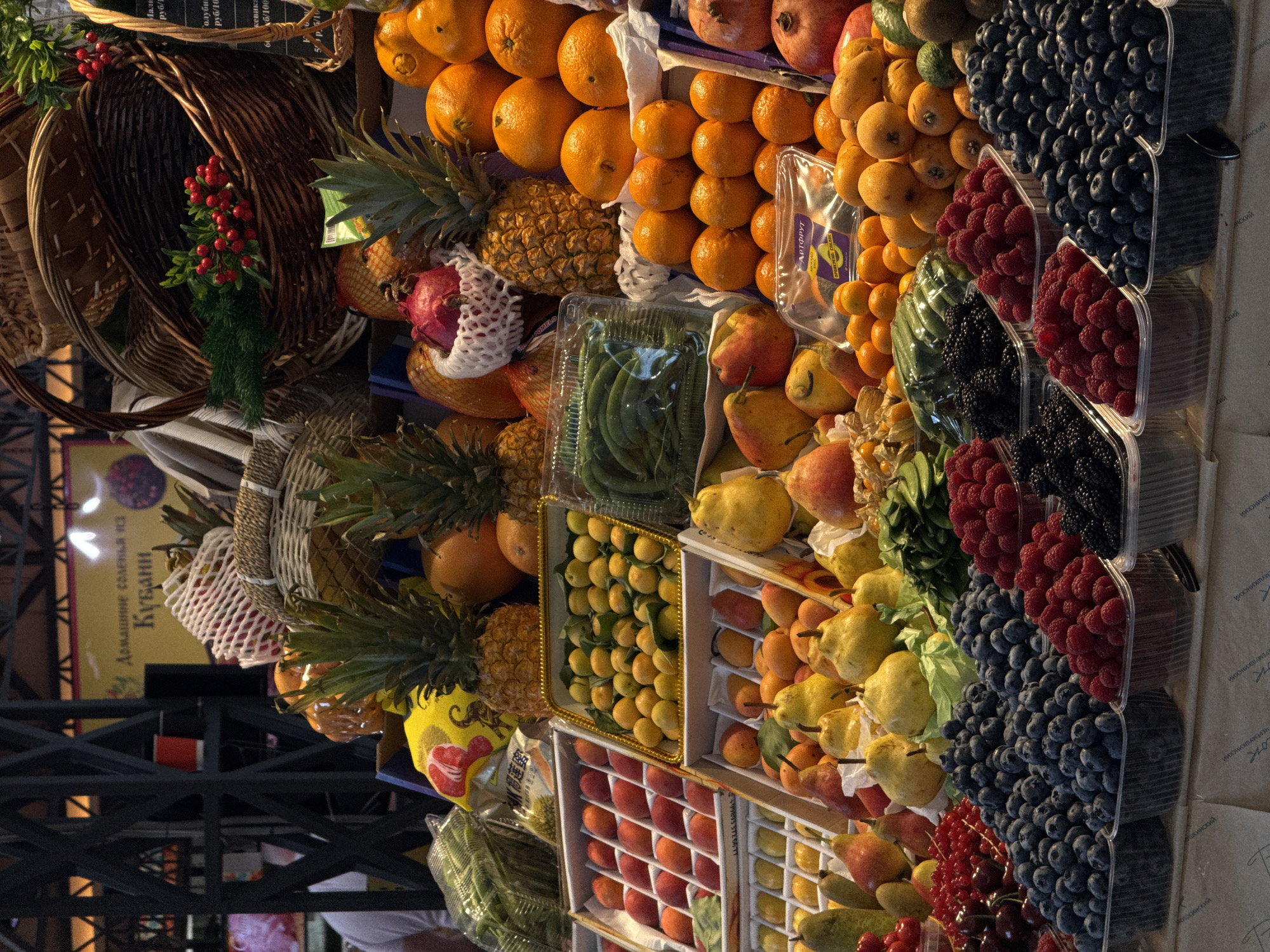 A detailed display of fresh fruits and berries is presented. The array includes oranges, mandarins, pineapples, pomegranates, loquats, pears, apricots, kiwis, blueberries, raspberries, blackberries, red currants, green beans, and persimmons. The fruits and berries are arranged in containers and baskets. The scene is brightly lit.
The background reveals posters in Russian, suggesting the location is Moscow, Russia. One sign reads "Domashnie Solen'ya Kubani" (Homemade Pickles of Kuban). Another sign appears to be an advertisement for "Artfrut" with pricing details, indicating a commercial environment.