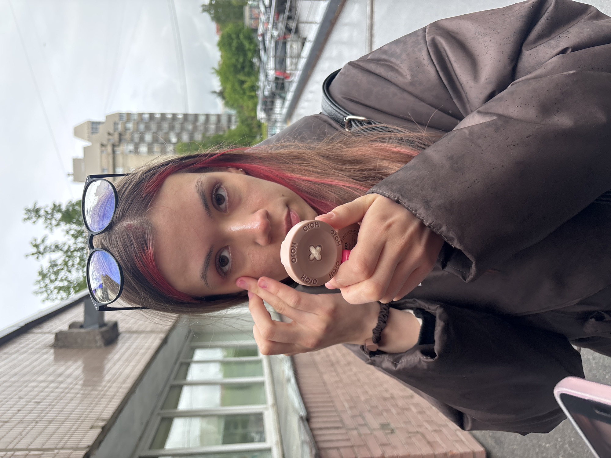 A young woman with brown hair and pink highlights, wearing glasses on top of her head, is applying makeup to her face outdoors. She is holding a small, circular makeup compact in her right hand, labeled "HOJO." Her left hand is raised to her face, near her cheekbone, as if she is applying the makeup. She is wearing a dark brown, possibly rain-resistant, jacket. In the background, there are trees and a multi-story building. The scene appears to be in Saint Petersburg, Russia, based on the style of the buildings and the wet weather conditions, which are typical for the city.