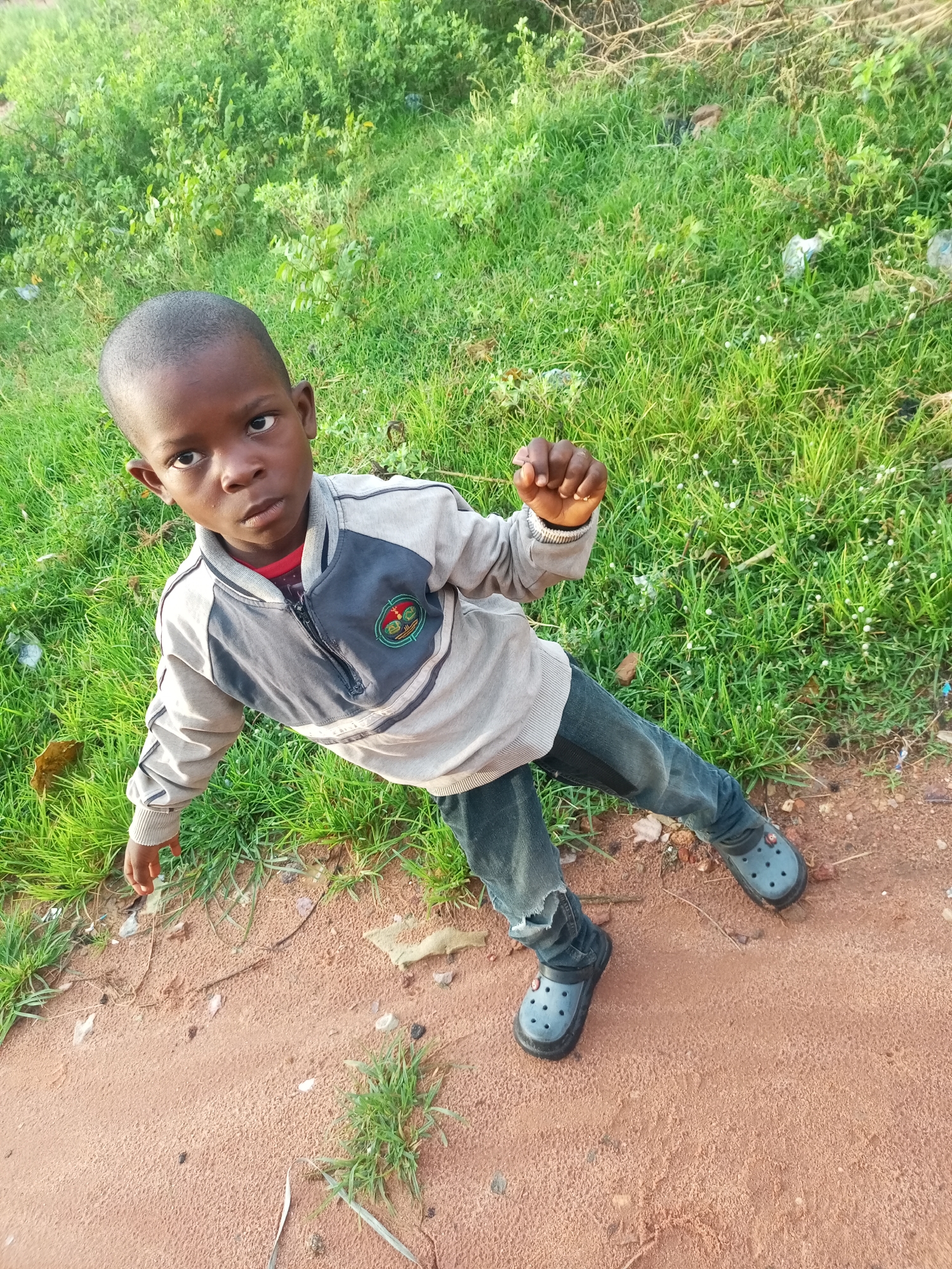 An eye-level, high-angle full shot shows a young Black boy standing on a dirt path with green grass to his left in Tempure, Nigeria. The boy is wearing a light-colored zip-up jacket with dark gray accents on the shoulders and chest, a red shirt underneath, blue jeans with rips at the knees, and blue Croc-like shoes. He has short, dark hair and is looking towards the camera with a neutral expression. His right arm is bent at the elbow with his fist clenched, while his left arm is extended downwards. The background consists of dense, green grass and some scattered debris, suggesting an outdoor, possibly undeveloped area within the city. The lighting suggests it is daytime, with natural light illuminating the scene.
