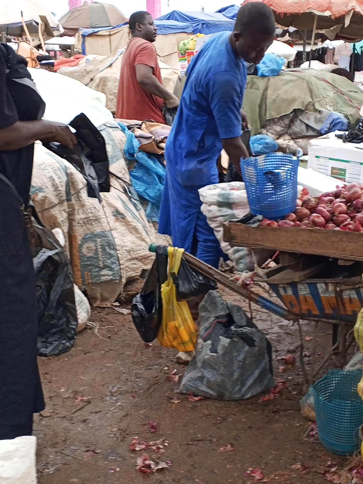 A man in a blue shirt and pants is standing at a market stall in Lokoja, Nigeria, and is sorting red onions. He is standing next to a wooden cart filled with onions, and a blue basket is placed on top of the cart. The man is also holding a black plastic bag. The ground is muddy and littered with onion skins. In the background, other market stalls and people are visible. The market appears to be an outdoor setting, with various goods displayed under awnings and tents. The overall impression is one of a bustling local marketplace.