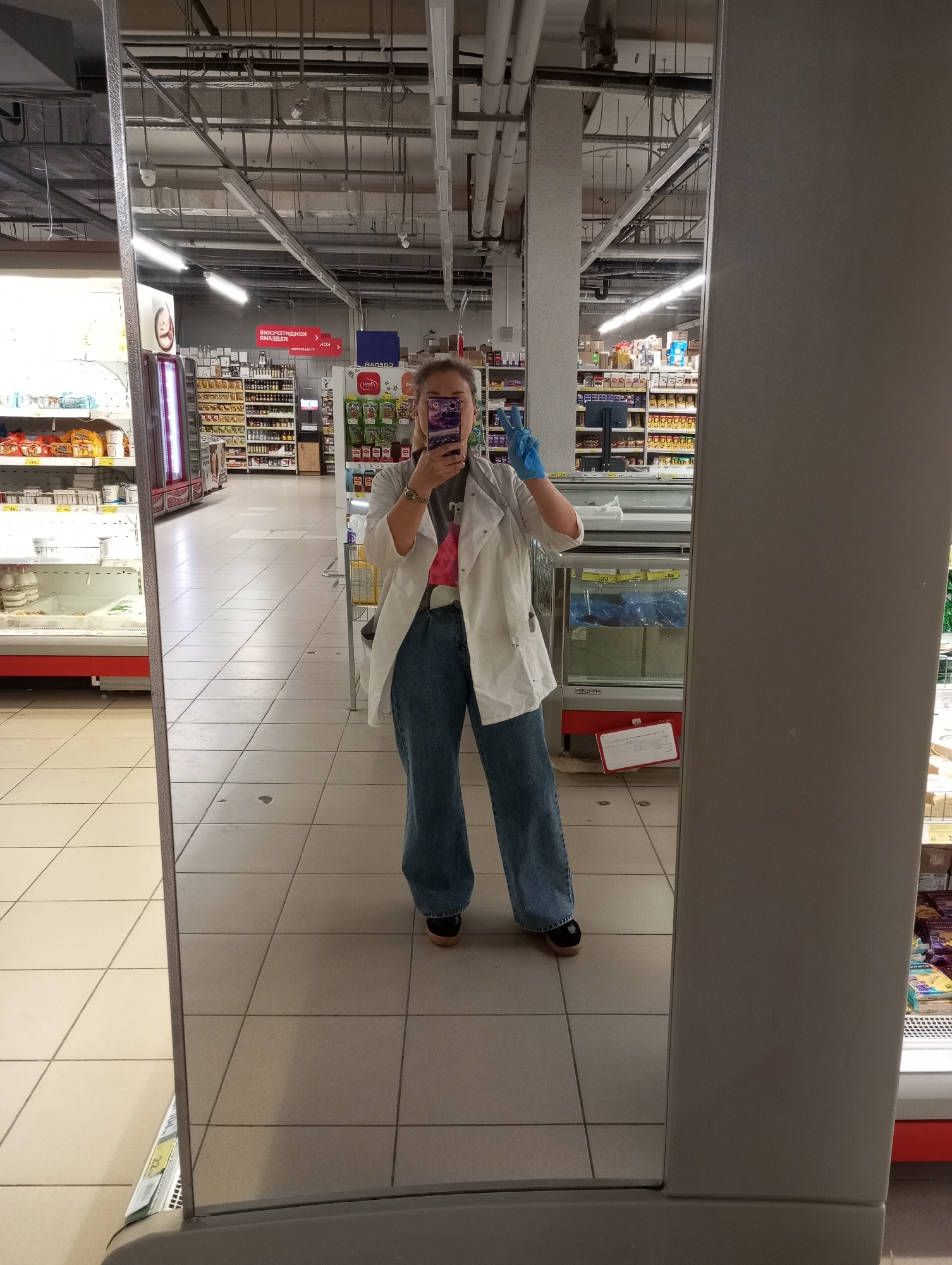 A full-body shot of a woman is captured in a mirror reflection, positioned within a supermarket setting, likely in Kamensk-Uralsky, Russia. The woman wears a white lab coat over a casual outfit, jeans, and dark shoes, along with blue gloves on one hand. She holds a phone in front of her face, presumably taking a selfie. The background shows typical supermarket aisles with shelves stocked with various products. The architecture includes overhead pipes and lighting fixtures common in such commercial spaces. A refrigerated display case is visible to the right of the woman in the reflection. The tiled floor indicates the interior of the store.