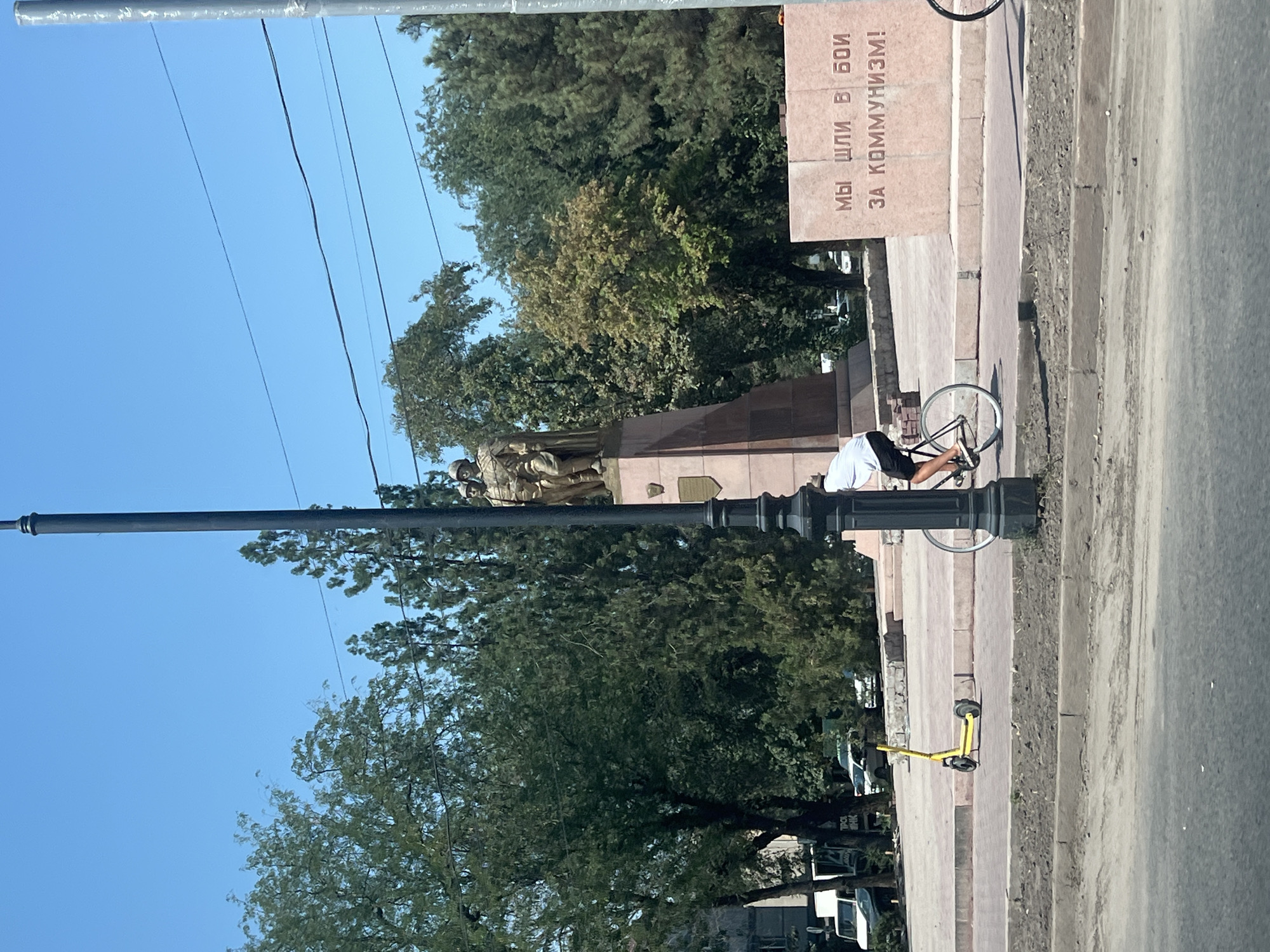 The image depicts a street scene in Bishkek, Kyrgyzstan. In the foreground, a cyclist is riding a bicycle on a paved path. To the left of the cyclist, a yellow electric scooter is parked. Behind the scooter, a statue of two figures stands on a pedestal. To the right of the statue, a large stone monument with Cyrillic lettering is visible. The background features trees and a clear blue sky. The scene appears to be a public space with pedestrian walkways and a road.