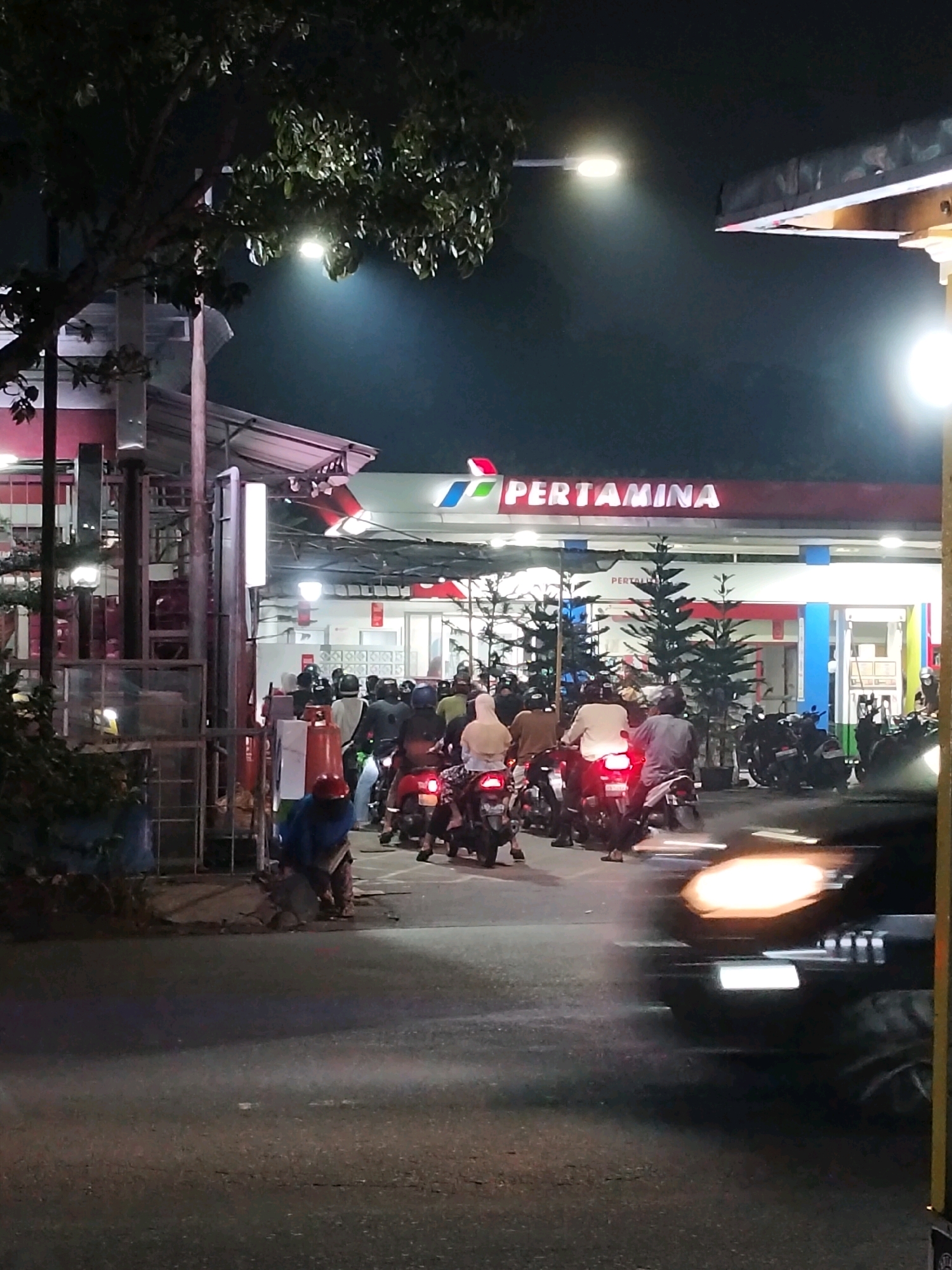 The image depicts a nighttime scene at a Pertamina gas station in Pontianak, Indonesia. A queue of motorcycles is lined up, indicating a possible rush or shortage. Several individuals are on the motorcycles, some wearing helmets, while others are standing or walking near their vehicles. The gas station features its characteristic red and white branding, with bright lights illuminating the forecourt. Trees are visible, and a street light casts a glow on the dark sky. A black car is captured in motion in the foreground, its headlights creating a blur. The ground appears to be asphalt, with some debris or unevenness visible. The overall atmosphere suggests a busy and somewhat chaotic environment.