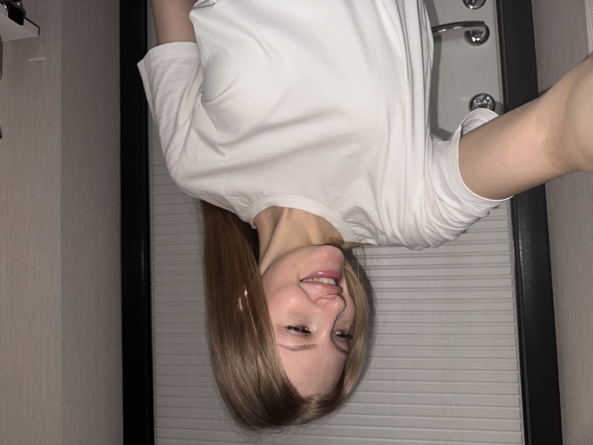 A blonde-haired woman is smiling while taking a selfie indoors, standing in front of a door with horizontal slats. The door has a dark frame and silver hardware. The wall to the left is beige with a subtle texture. The city of Perm, Russia, cannot be confirmed from this image.