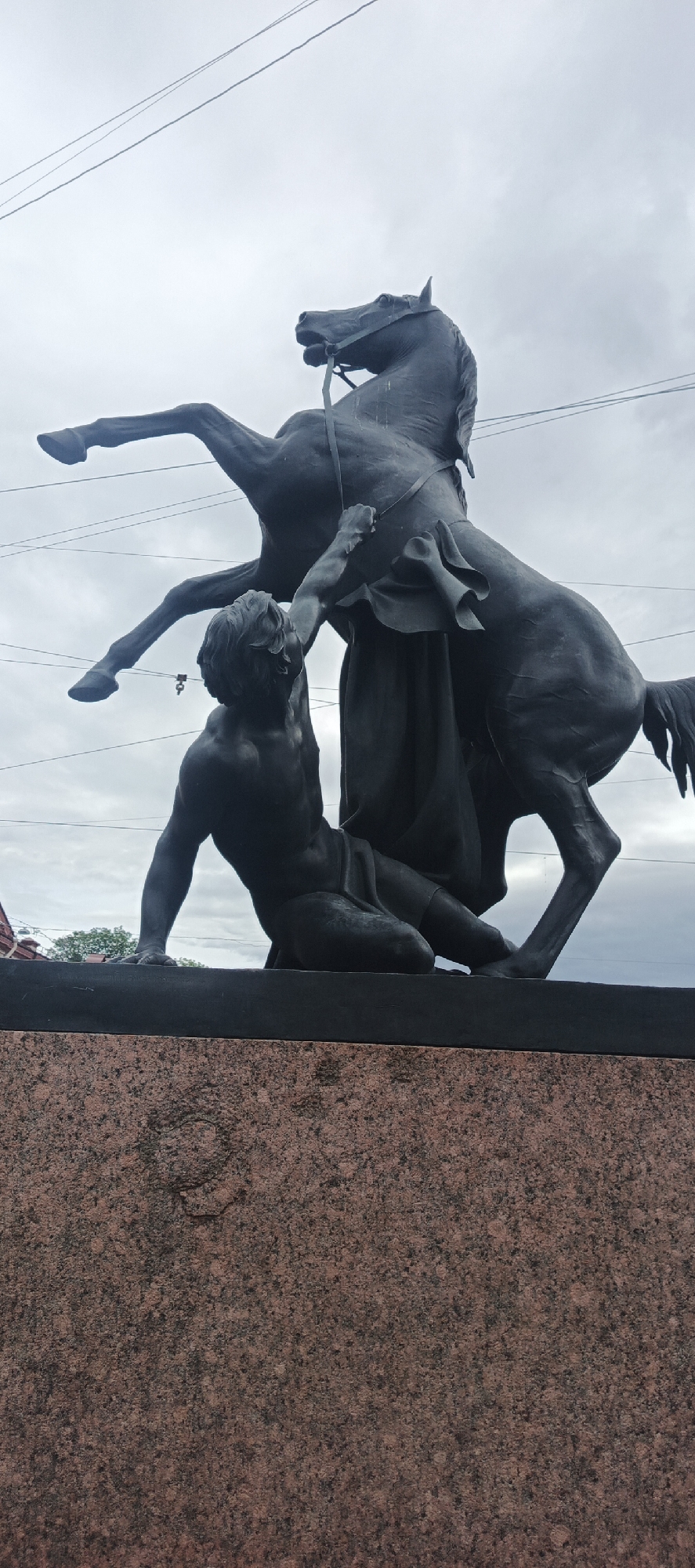This is a low-angle, full shot of a monumental bronze sculpture depicting a man wrestling with a rearing horse. The man is muscular and appears to be struggling to control the powerful animal. He is kneeling on a dark, flat surface, with his left arm raised, gripping the horse's bridle, while his right arm is extended upwards, grasping the horse's mane or forelock. The horse is depicted mid-rearing, with its front legs off the ground and its head turned slightly to its left. A dark, flowing cloth or garment is draped over the horse's back.
The sculpture is mounted on a base made of speckled pink and black granite. Above the sculpture, a cloudy, overcast sky is visible, with several thin, dark electrical wires crisscrossing the frame. In the far background, the tops of trees and a building with red accents can be discerned, suggesting an urban setting. The overall mood of the sculpture is one of intense struggle and raw power. The lighting suggests it is daytime, but the cloudy sky indicates overcast weather.
The location is identified as Saint Petersburg, Russia. There is no visible text within the image.