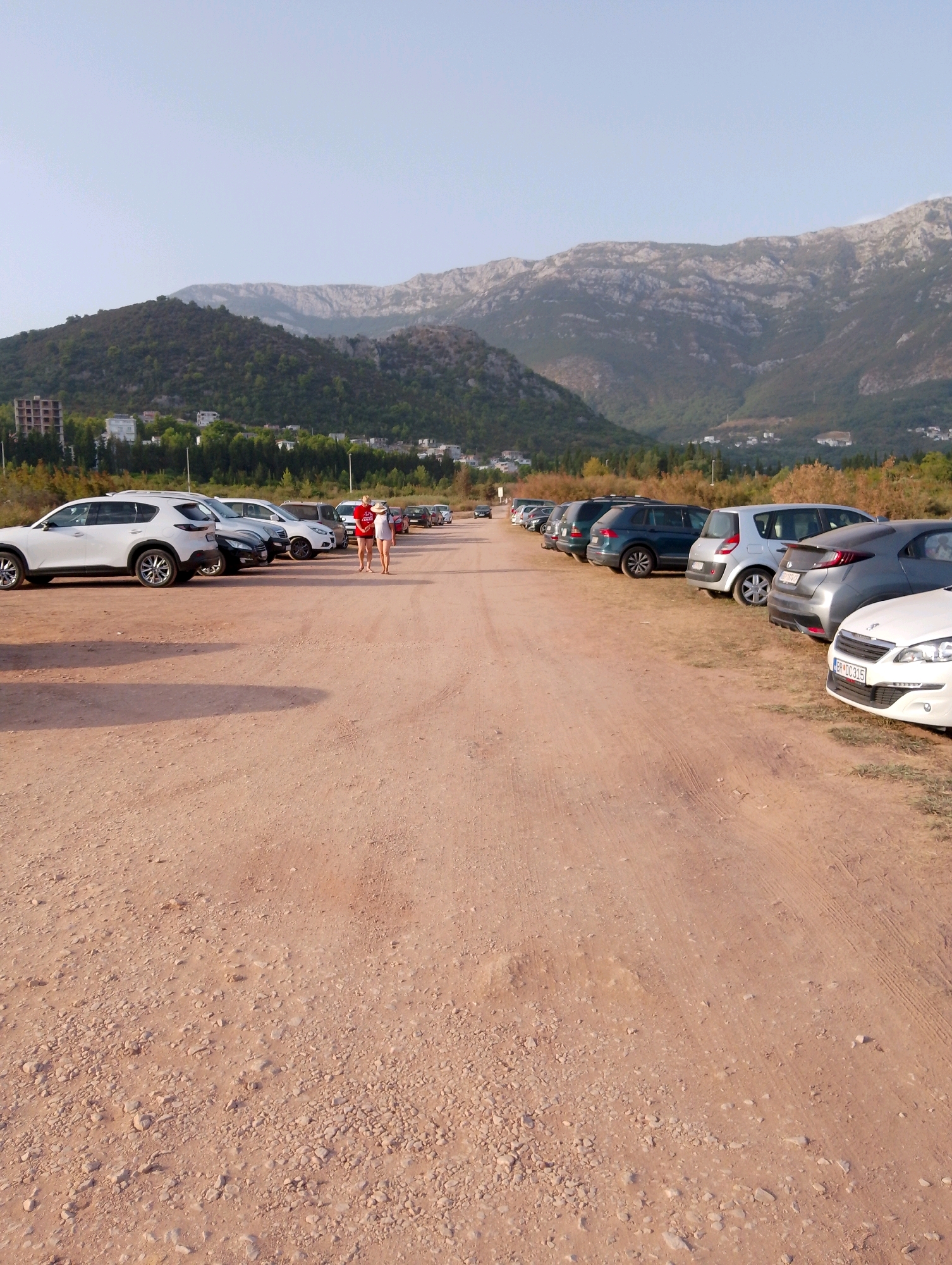 The image depicts a dirt parking lot in Zagrađe, Montenegro, with mountains in the background. Several cars are parked in a line on the right side of the lot. Two individuals, a woman in a white swimsuit and a straw hat and a man in a red t-shirt and shorts, are standing and interacting in the middle of the parking lot, to the left of the parked cars. The ground is a light brown, dusty earth with scattered small stones. Trees and buildings are visible on the lower slopes of the mountains, with a clear sky above.