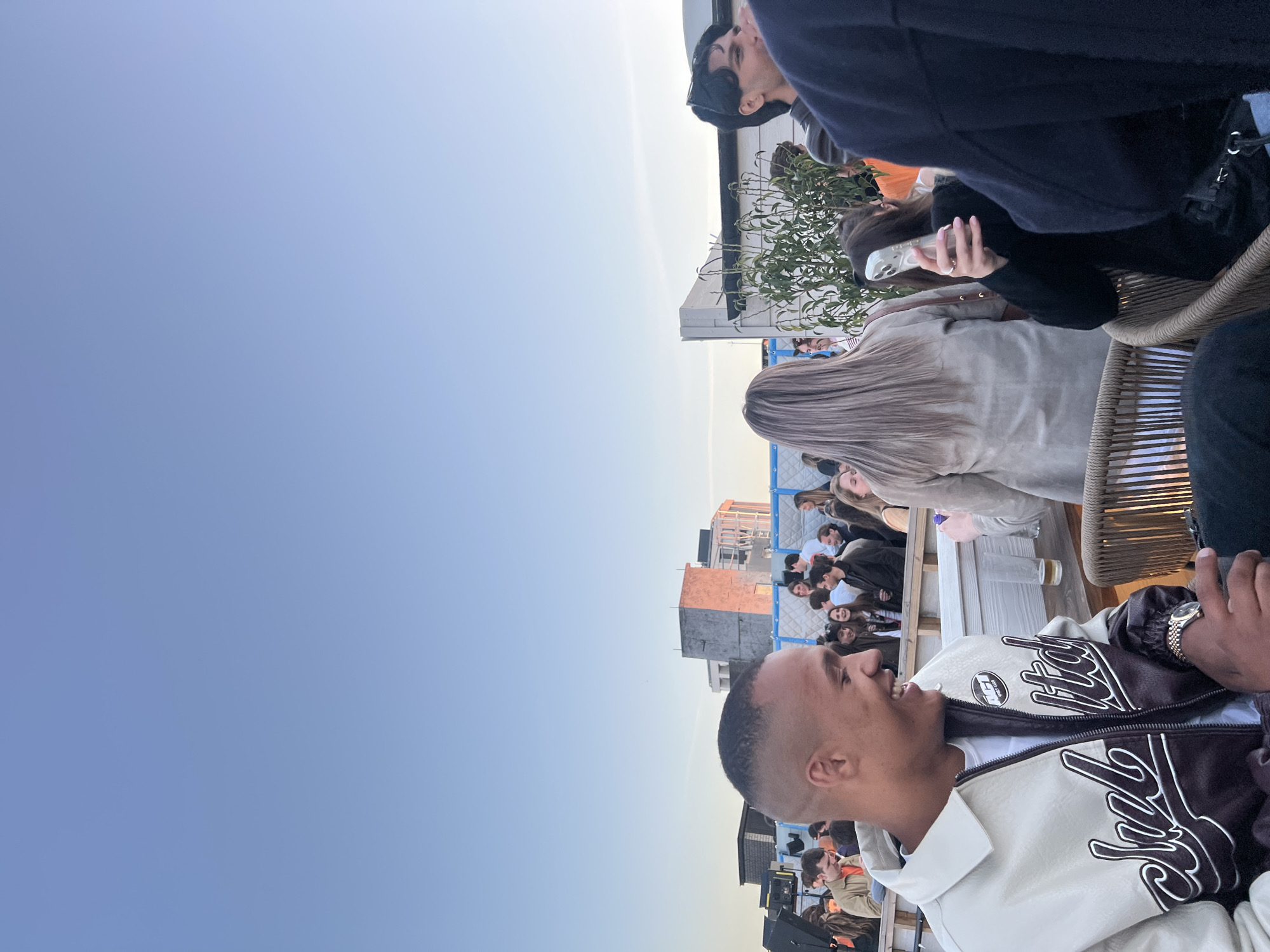 A daytime rooftop gathering in London, United Kingdom. People are seated at tables, socializing and enjoying drinks. Buildings are visible in the background. The sky is clear and blue.