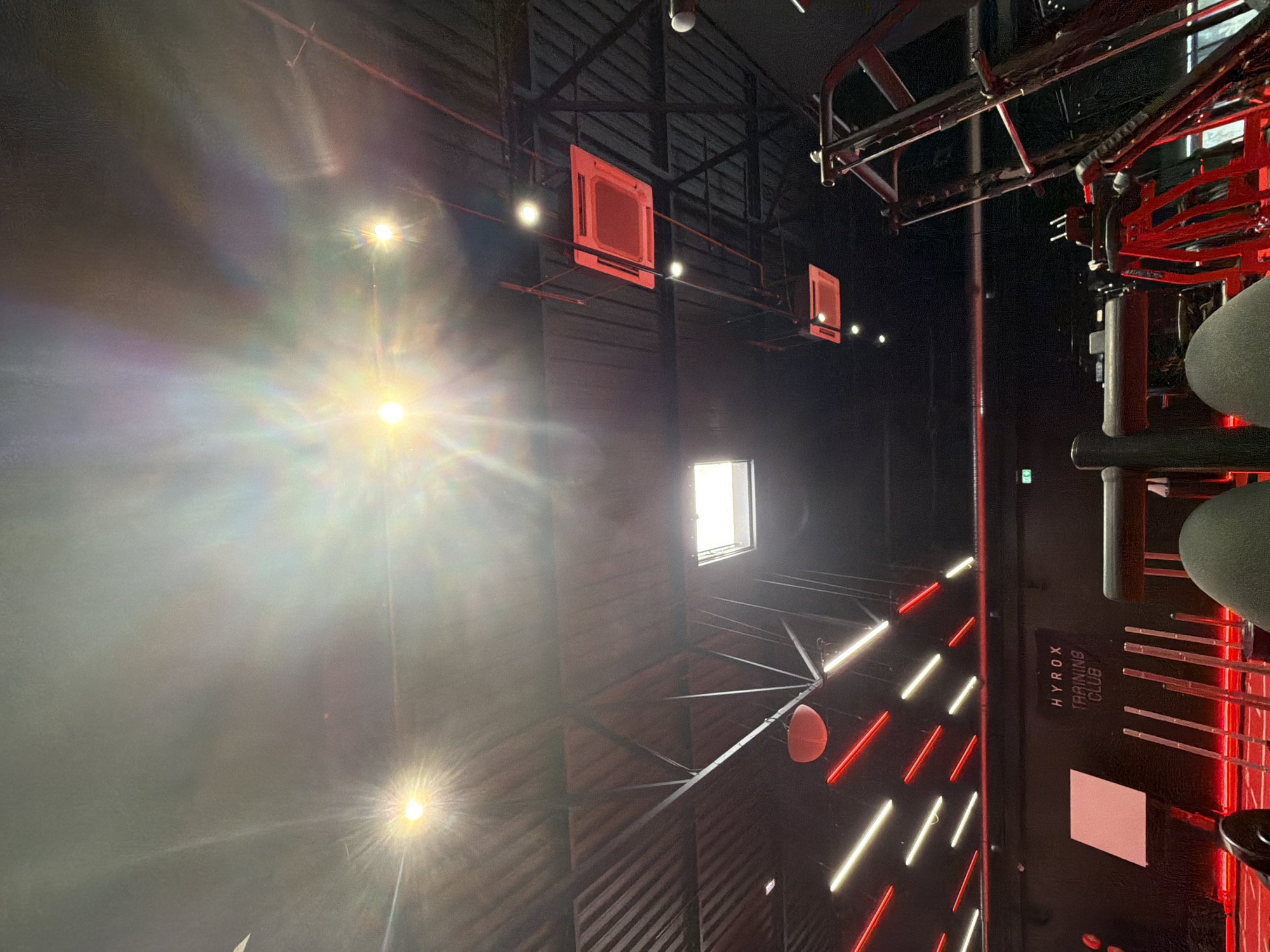 This image depicts the interior of a gym, specifically the HYROX TRAINING CLUB, located in Strasbourg, France. The viewpoint is from a low angle, looking up towards the ceiling which features a dark, ribbed texture with exposed metal beams and ductwork. Several bright lights are visible, including two prominent circular lights casting lens flares and a large rectangular skylight. Red and white LED lighting strips are mounted on the ceiling, creating a vibrant, modern atmosphere.
In the foreground, parts of gym equipment are visible, including a weight rack and what appears to be a leg press machine. The presence of two human legs, clad in dark grey, suggests a person is seated on or interacting with one of the machines. A banner with the HYROX TRAINING CLUB logo is mounted on a black wall in the background. The overall environment suggests a busy, well-equipped fitness facility.