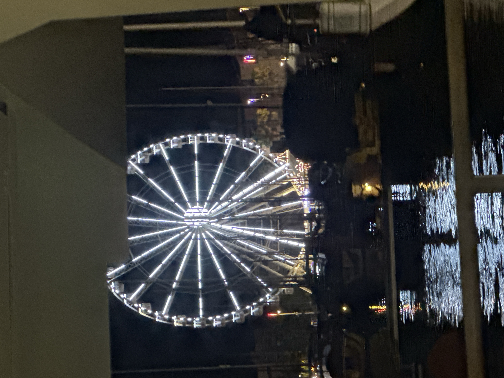 The image shows a Ferris wheel illuminated at night, likely in a city or entertainment district. The Ferris wheel is the central and most prominent object, with its spokes and carriages clearly defined by bright white lights. It is set against a dark sky, suggesting it is nighttime. In the foreground, there is water, possibly a harbor or river, with reflections of lights on its surface. Some boats or structures are visible in the water. A dark, silhouetted shape, possibly people or structures, is present in the middle ground, obscuring part of the view of the Ferris wheel and the background. The overall impression is a lively evening scene, possibly at a fair or waterfront attraction. The question of the location in the city of Agde, France, is not directly answerable from the visual information provided in the image alone.