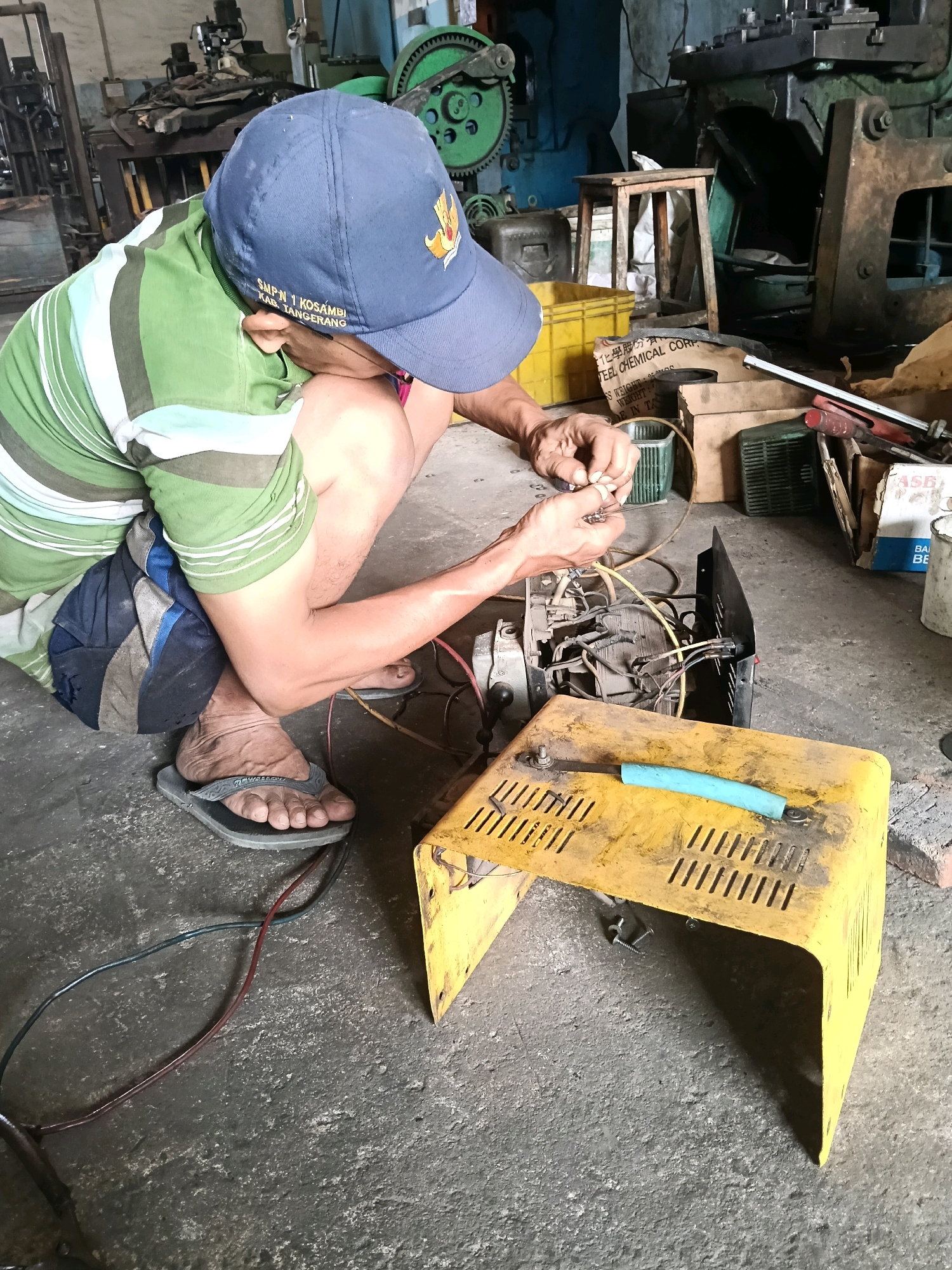 A man wearing a green and white striped shirt, blue baseball cap, and flip-flops is crouched on a concrete floor, working on electrical components of a black device. Wires and tools are scattered around the device, which sits on a yellow metal stand. The background reveals a workshop environment with various machinery and storage containers, including a large green gear. The man appears to be in the city of Belimbing, Indonesia, given the text on his cap which reads "SMPN 1 KOSAMBI KAB. TANGERANG".