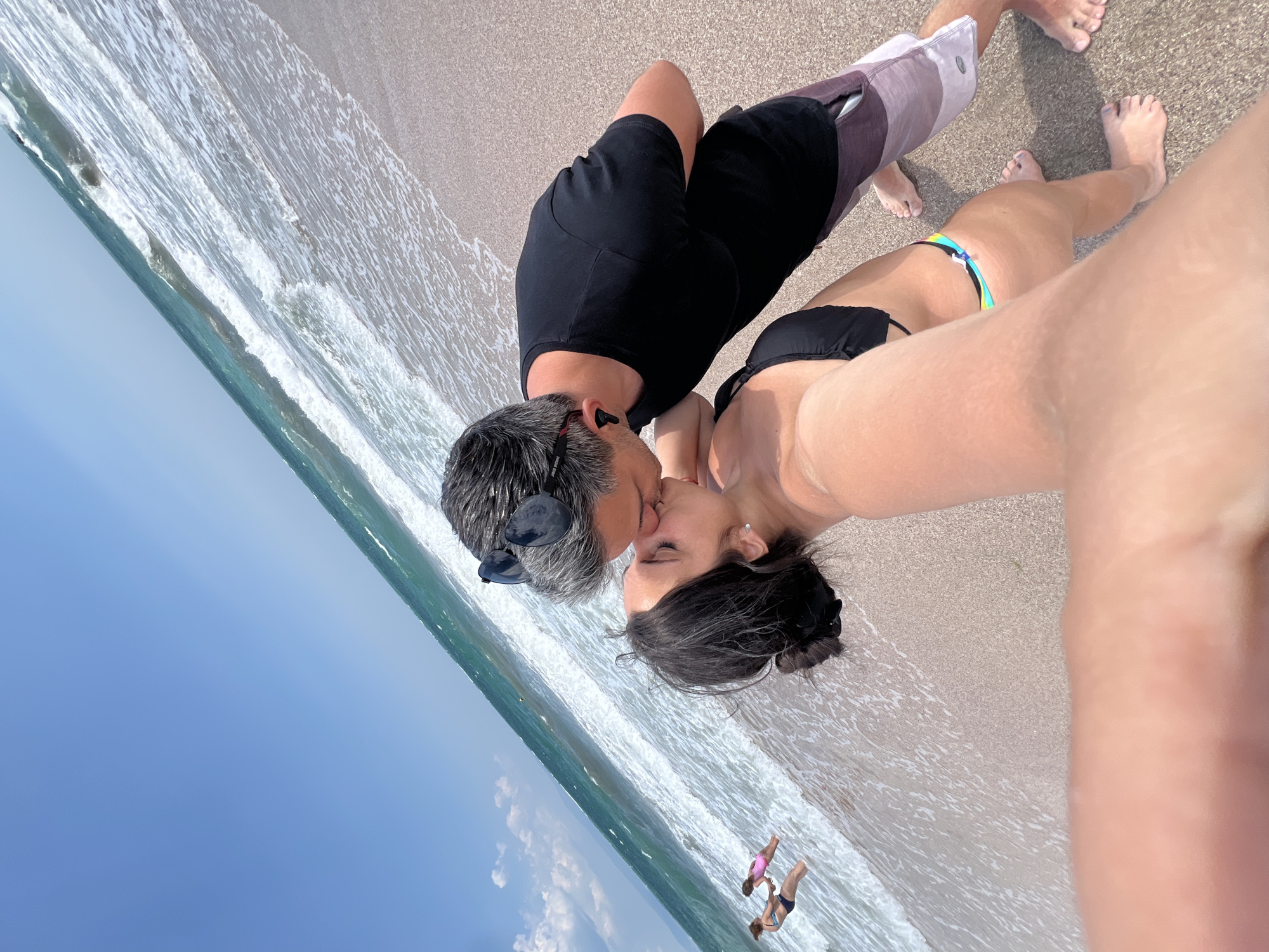 A high-angle, selfie-style photograph captures a couple kissing on a beach in Pomorie, Bulgaria. The couple is positioned in the foreground, with the woman in a black bikini and the man in a black t-shirt and board shorts. The ocean, with its characteristic foamy waves, stretches out behind them. In the background, two young girls in swimwear can be seen wading in the shallow water, their figures small against the vastness of the sea and sky. The beach sand is a light, sandy brown, and the water is a clear, turquoise blue. The sky is a bright, cloudless blue, with a hint of haze in the distance. The overall impression is one of a romantic and idyllic beach scene on a sunny day.
