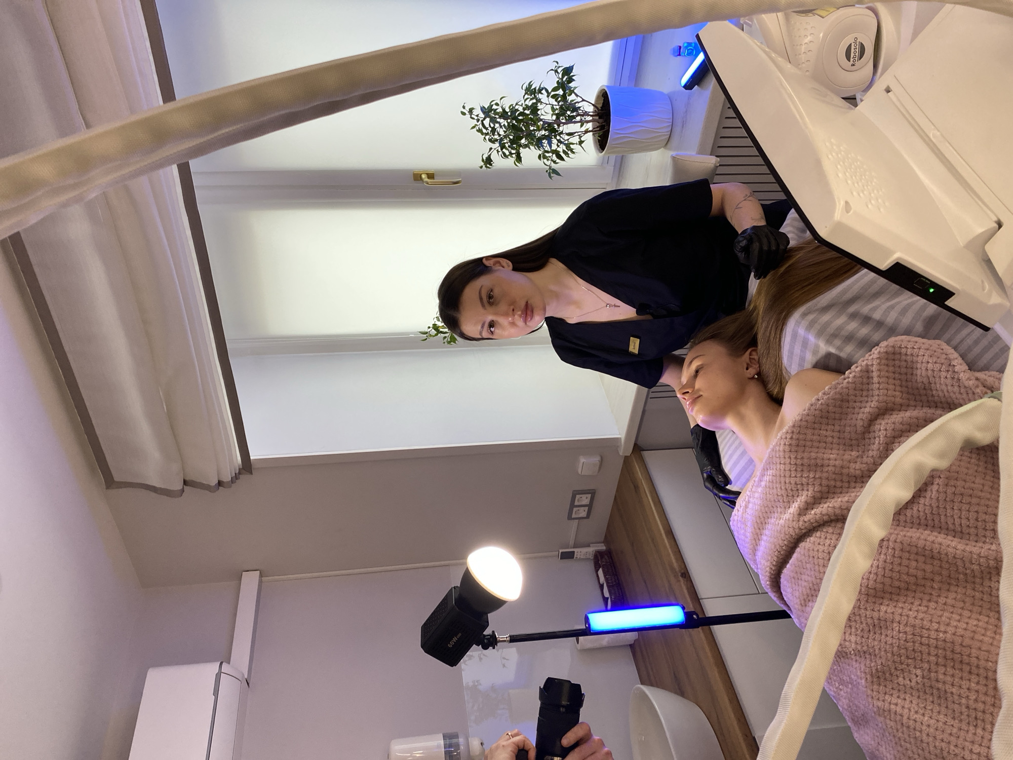 This image captures an indoor scene within what appears to be a modern beauty salon or aesthetic clinic, likely in Votkinsk, Russia. The perspective is tilted, looking down at the subjects.
In the foreground, a female practitioner stands, wearing a black long-sleeved top and black gloves, looking directly at the camera with a neutral expression. A small yellow badge is pinned to her top. She has dark, long hair. Adjacent to her, a female client lies reclined on a treatment bed, covered by a soft, pink textured blanket or robe. Her eyes are closed, and she appears relaxed, undergoing a procedure. Her light brown hair is spread around her head.
A large, white, futuristic-looking machine is positioned over the client's upper body, extending from above her head. The brand name "Robosolo" is visible on a part of this equipment. Hoses or tubes run from the machine, suggesting a targeted treatment.
The room is brightly lit, with natural light entering through a large window covered by sheer, light-colored curtains. A small potted plant sits on the windowsill. Artificial lighting enhances the scene; a circular LED light marked "60W" is mounted on a stand in the lower left, and a blue light bar is visible on another stand nearby. The presence of these professional lights, along with a camera lens and body held by unseen hands in the very bottom left corner, suggests that the treatment is being documented, possibly for promotional purposes.
The walls are white, and a section of a wooden counter or shelf is visible to the right. Electrical outlets are also seen on the wall. The overall ambiance is clean, professional, and well-equipped for aesthetic procedures.