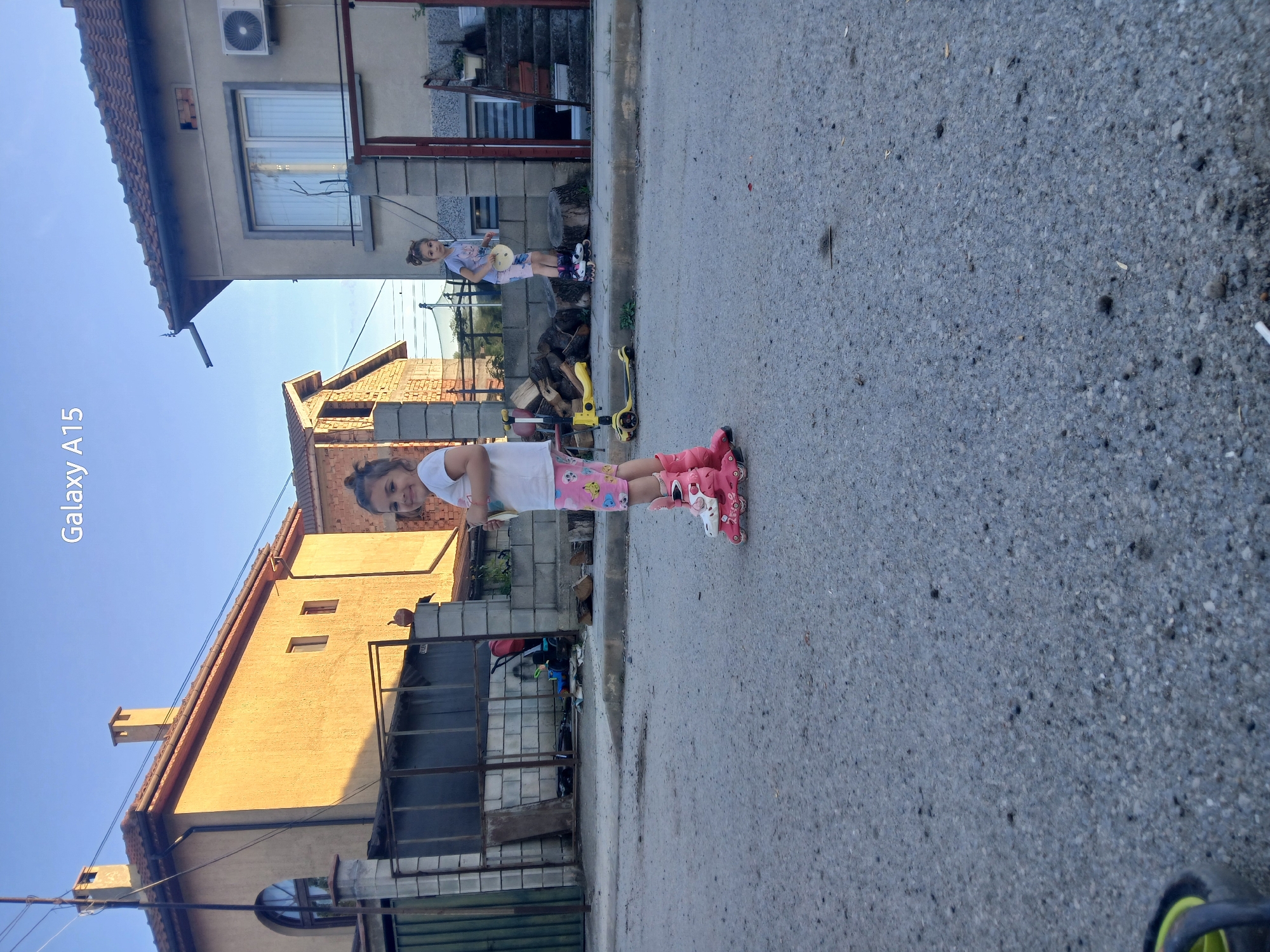 Two young girls are on a residential street in Obrochishte, Bulgaria. One girl is wearing pink roller skates, a white t-shirt, and patterned shorts. She is standing in the street and facing the camera. The other girl is slightly further away, near a building, holding a round yellow object, and also appears to be wearing roller skates. The street is asphalt, and the background includes houses, a yellow scooter, and some stacked wood. The weather appears sunny and clear. The image is taken from a mobile phone, with the "Galaxy A15" watermark present on the left side.
