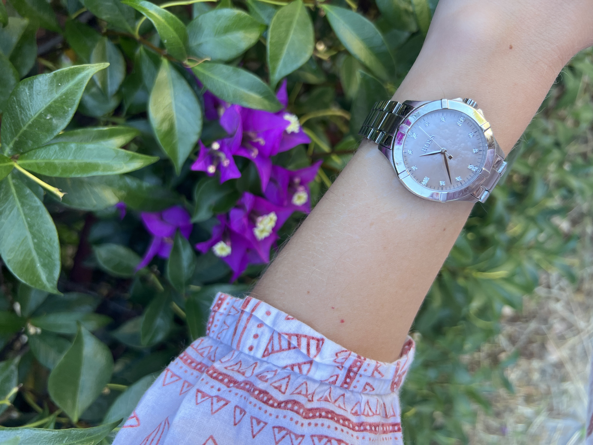 A person's arm, wearing a silver watch with a light pink mother-of-pearl face and diamond markers, is in the foreground, angled towards the right. The watch is positioned on the wrist, which is attached to the arm. The arm is lightly tanned and has sparse light-colored hair. The person is wearing a white blouse with a red geometric pattern on the cuff. The background is a dense green bush with vibrant purple bougainvillea flowers. The scene is outdoors, with natural lighting. There are no buildings, streets, or any specific landmarks visible that would indicate a location in Budva, Montenegro.