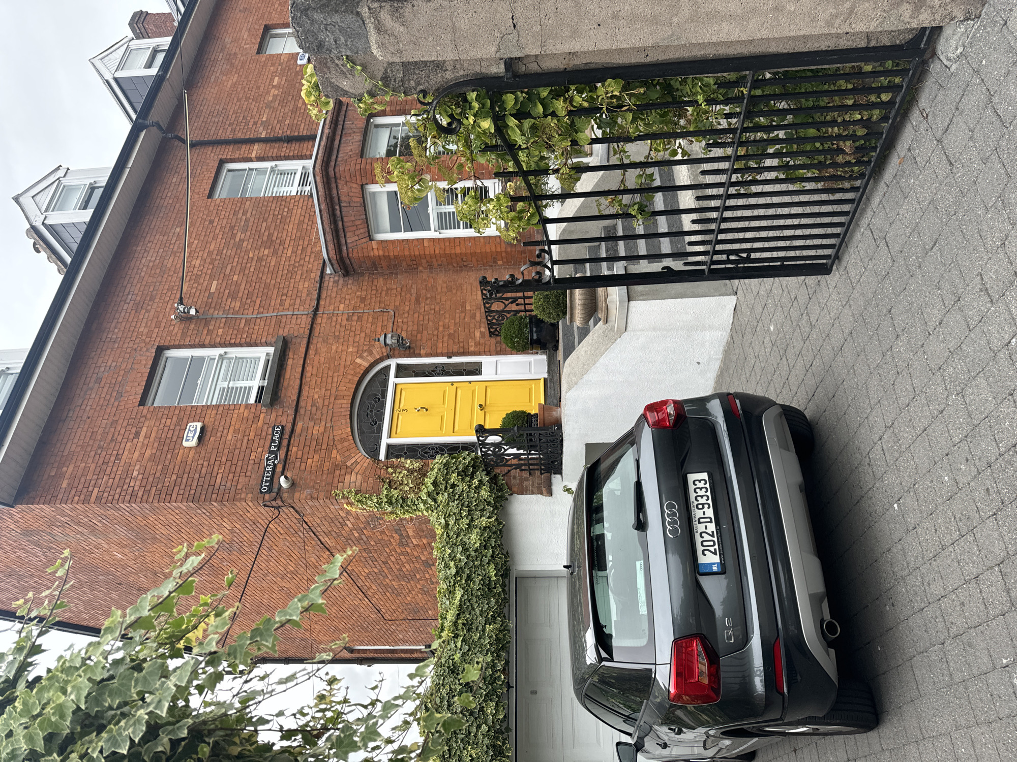 A dark grey Audi Q2 is parked on a paved driveway in front of a red brick house with a bright yellow front door. The house has white window frames and a white garage door. Ivy covers part of the house facade and a stone wall. A black metal gate is partially open to the right of the image. The license plate on the Audi reads "202-D-9333". A sign above the entrance reads "OTTERAN PLACE". The scene is likely in Waterford City, Ireland, given the license plate format and the presence of a sign indicating a street name. The sky is overcast.