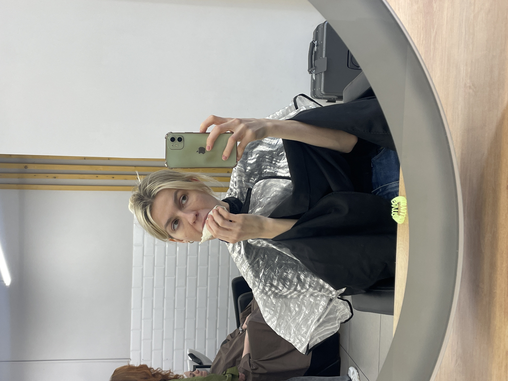 A woman with blond hair is visible in a mirror, holding a smartphone and eating while wearing a stylist's cape. A white tiled wall is visible behind her. Other customers are in the room, as seen in the mirror's reflection. A grey hard-shell suitcase sits on a shelf behind her. Based on the interior design, it is possible that the image was taken in a salon in Moscow, Russia.