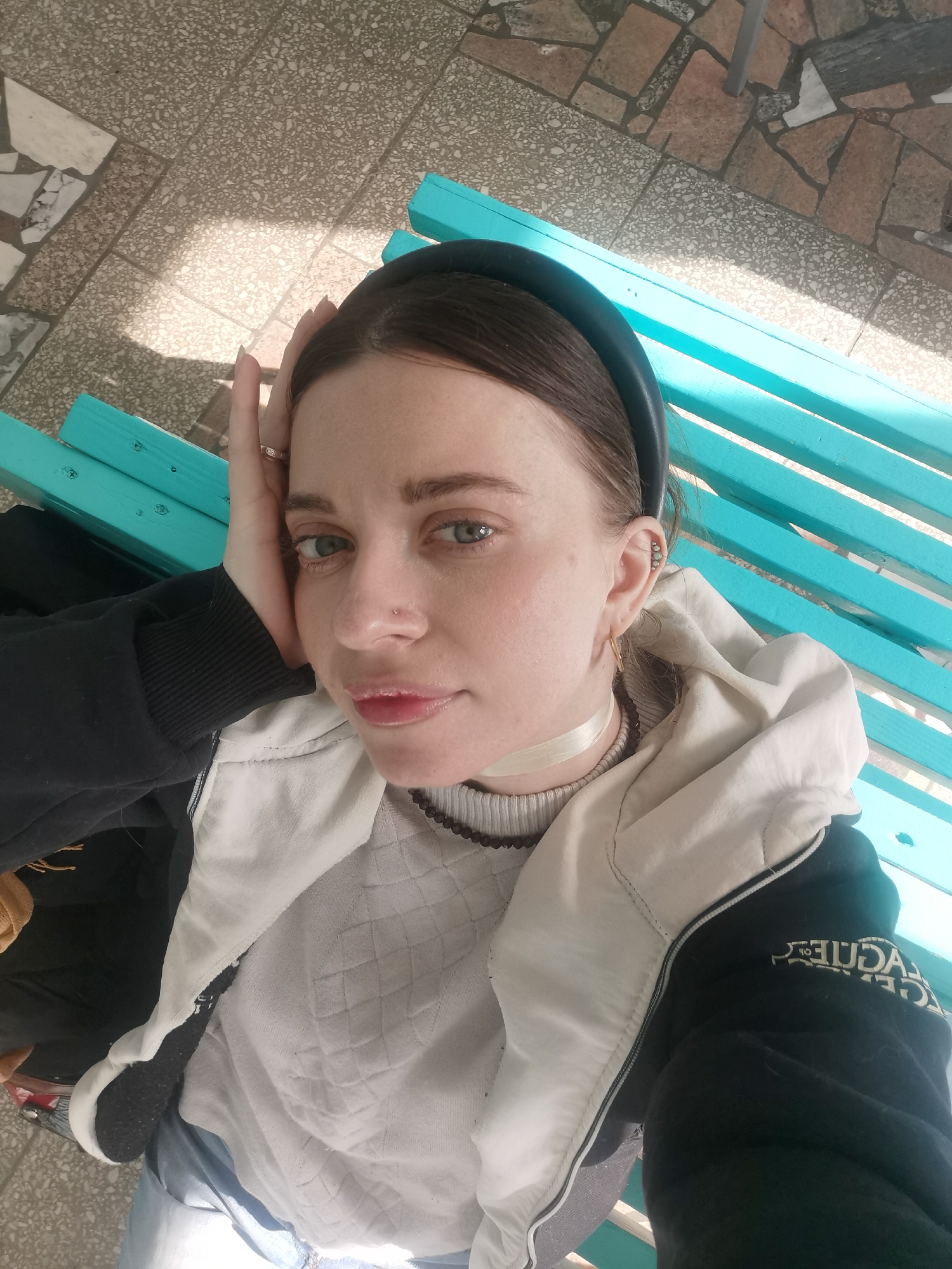 A young woman is depicted in a close-up, high-angle shot, seemingly taking a selfie while reclining on a bright turquoise slatted bench. She has dark hair held back by a black headband and looks directly at the camera with a neutral, relaxed expression. Her right hand, adorned with a ring, rests by her cheek. She wears a white quilted sweater or top, a white ribbon-like choker, and a black and beige/cream jacket/hoodie. A brown beaded necklace is partially visible around her neck. She has a small stud piercing on her nose and multiple piercings on her ear, including a hoop and several small studs along the helix.
The scene is outdoors, with strong sunlight casting shadows on the patterned paving stones that make up the ground. The weather appears sunny, indicating daytime. On the sleeve of her jacket, mirrored text is visible, spelling "LEAGUE OF LEGENDS". The background suggests an outdoor public space, possibly a park or patio area.