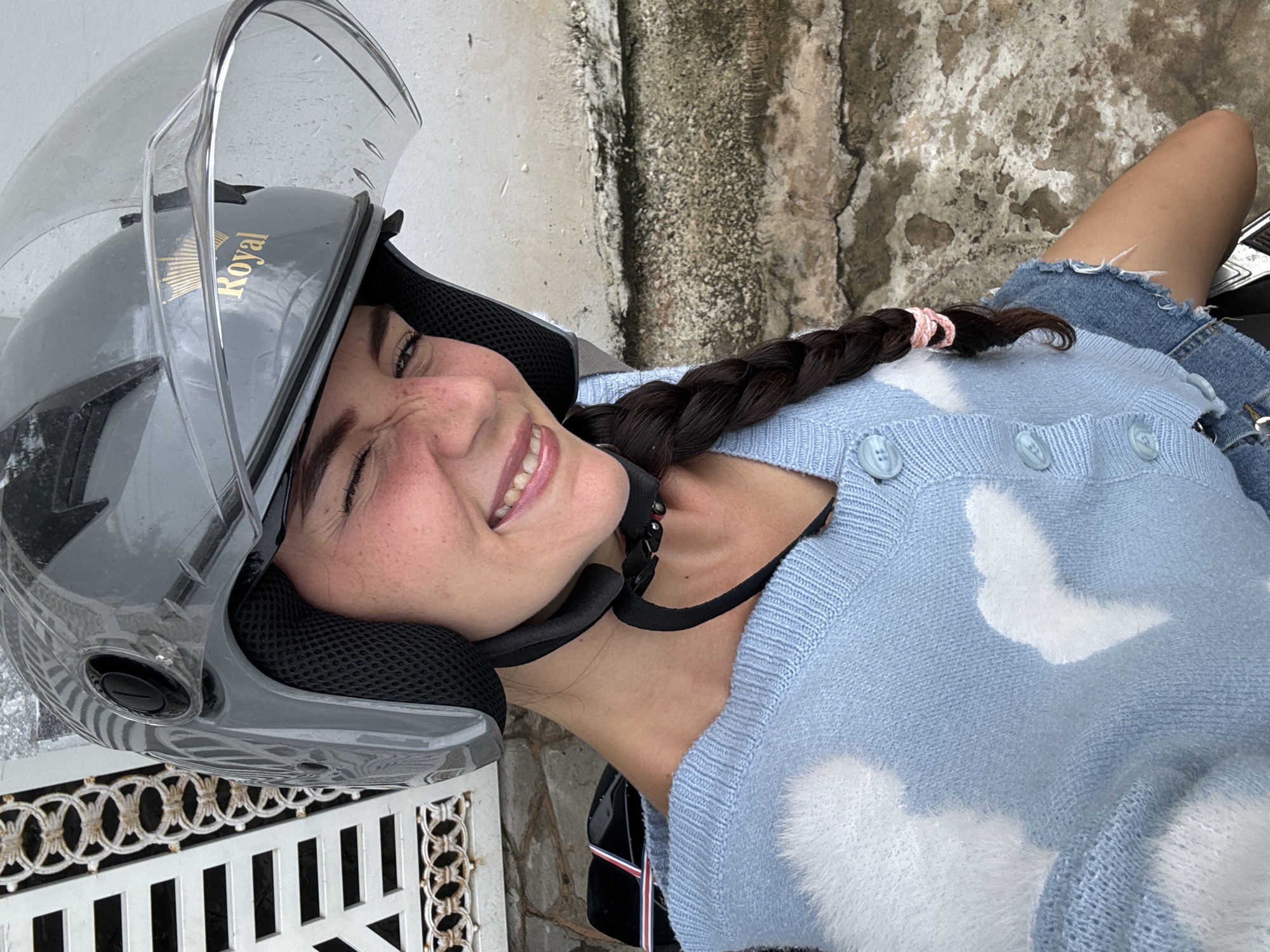 A young woman wearing a grey helmet with a clear visor and a light blue cardigan with white clouds is posing for a photo. Her hair is braided with a pink scrunchie. She is wearing a black shirt underneath the cardigan and a denim skirt. In the background, there is a textured wall and a decorative white metal fence. The location is likely Mũi Né, Vietnam.