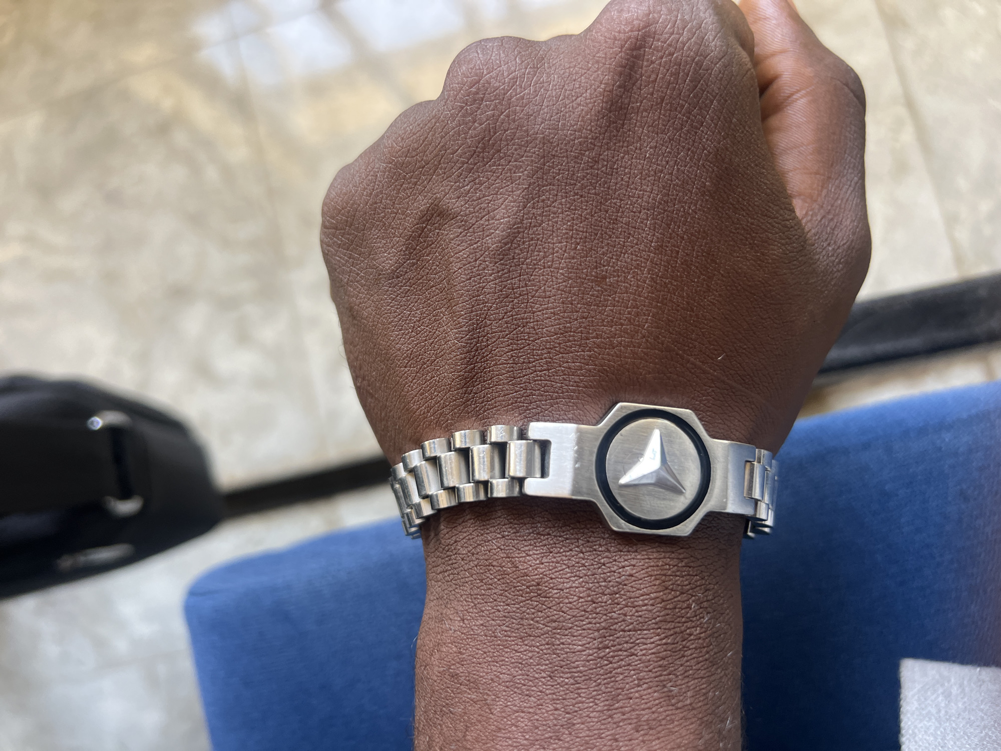 A close-up shot showcases a person's fist and wrist wearing a silver-tone metal bracelet featuring a prominent Mercedes-Benz logo at its center. The bracelet is comprised of interconnected rectangular links. The person's skin tone is dark. In the background, there is a partial view of a blue garment or upholstery and a black object, possibly a case or bag. The surface beneath appears to be a light-colored, possibly tiled floor. The location in the city of Jalingo, Nigeria is not discernible from the image.