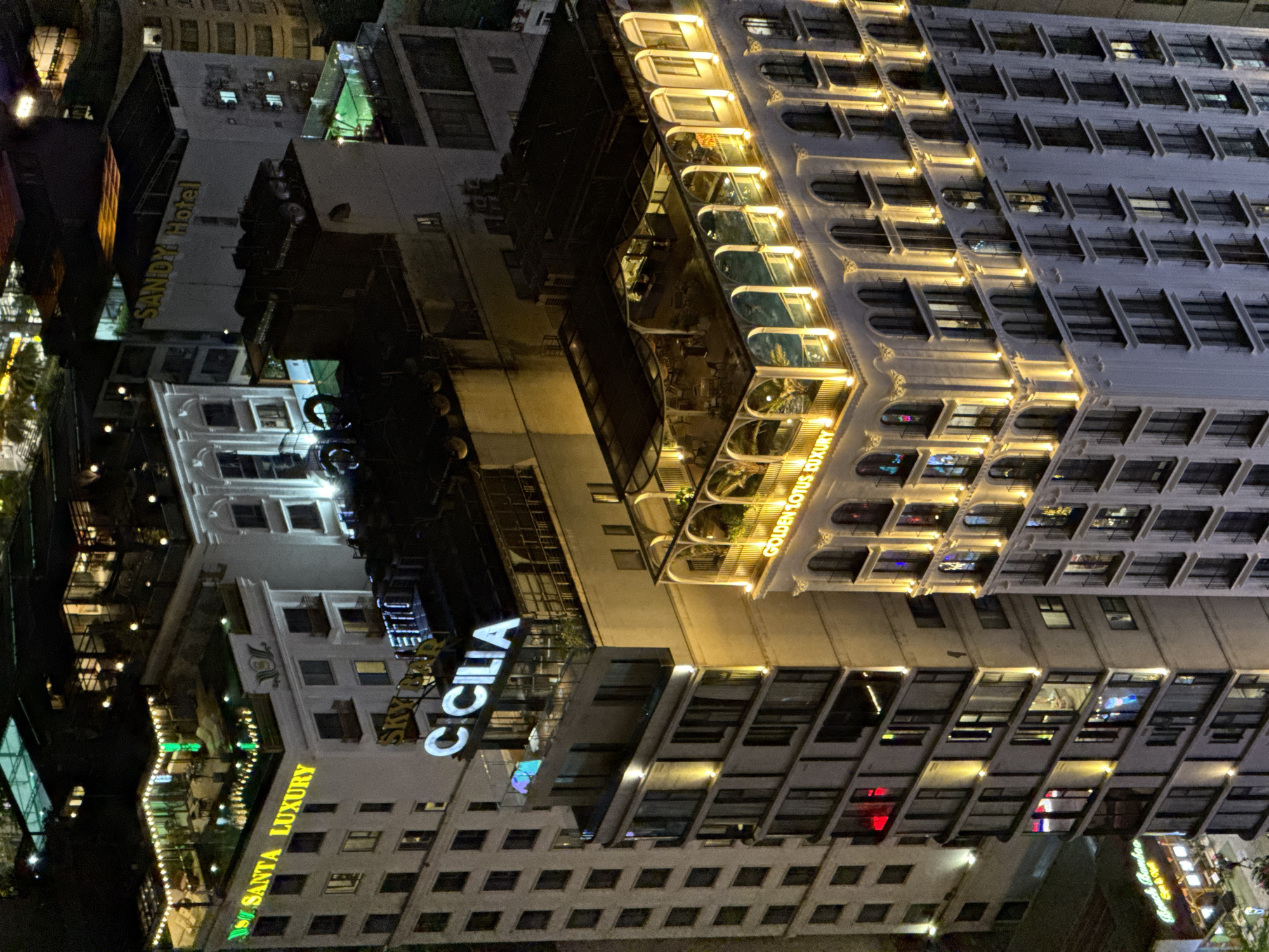 This media file captures a vibrant cityscape at night in Đà Nẵng, Vietnam, viewed from a high vantage point looking down upon a dense cluster of illuminated buildings. The scene is dominated by multiple multi-story structures, predominantly hotels or luxury apartment complexes, all brightly lit against the dark sky.
The most prominent building, positioned on the right, features intricate facade lighting with warm yellow tones that highlight its architectural details and numerous arched windows. Its lower levels prominently display the text "GOLDEN LOTUS LUXURY" and reveal an inviting outdoor terrace or lounge area with visible seating.
To the left of this structure, another building stands out with large, glowing white letters spelling "C CILIA." Further into the background and to the left, a green sign reads "SANTA LUXURY," while another building in the mid-ground features a yellow sign for "SANDY Hotel."
The buildings exhibit various architectural styles, from modern glass facades to more traditional designs with balconies and decorative elements. Many windows are illuminated, suggesting activity within the premises. The overall setting depicts a lively commercial or tourist district, bustling even after dark, creating a dynamic urban night scene. No people are distinctly visible, but the lights and visible outdoor seating imply human presence and activity within this part of Đà Nẵng.