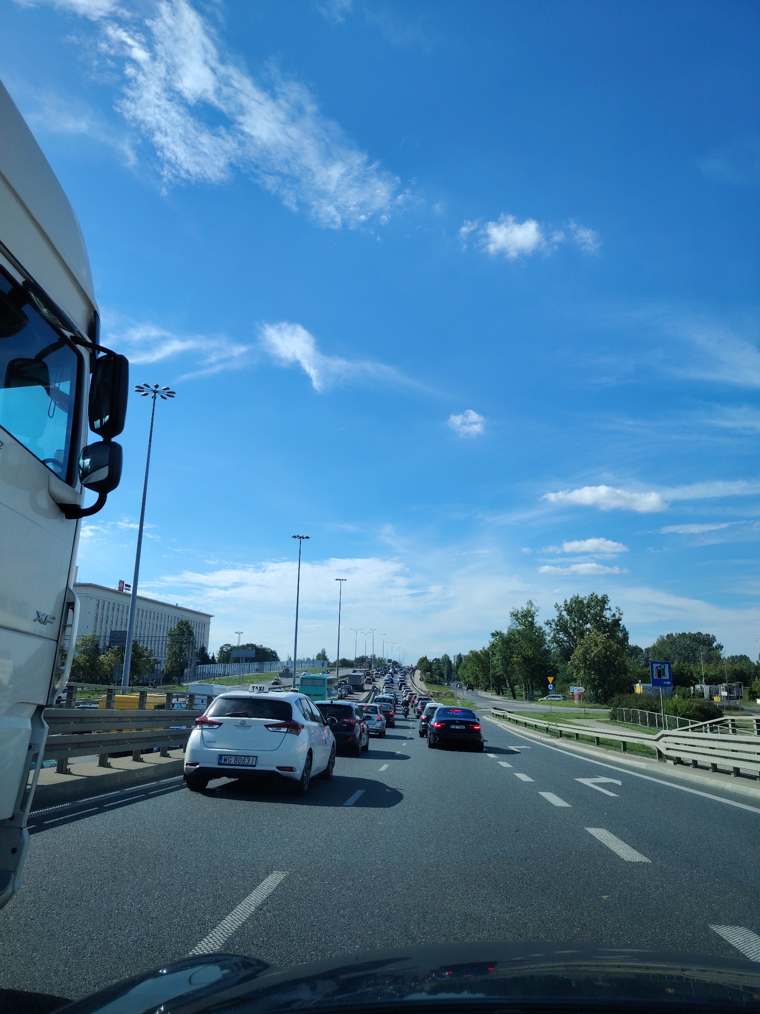 The image shows a traffic jam on a sunny day in Warsaw, Poland. The perspective is from inside a vehicle, looking forward. On the left, the side of a white truck is visible, with its side mirror and part of the cabin. The road is a multi-lane highway with a clear blue sky and scattered white clouds above. Various cars are seen in traffic, including a white hatchback with a license plate "WG 8063J" and a black sedan. Tall lampposts line the road. In the background, there is a large, light-colored building with many windows, possibly an office building or a public institution, with trees and greenery alongside the road. The traffic appears to be moving slowly or is at a standstill.