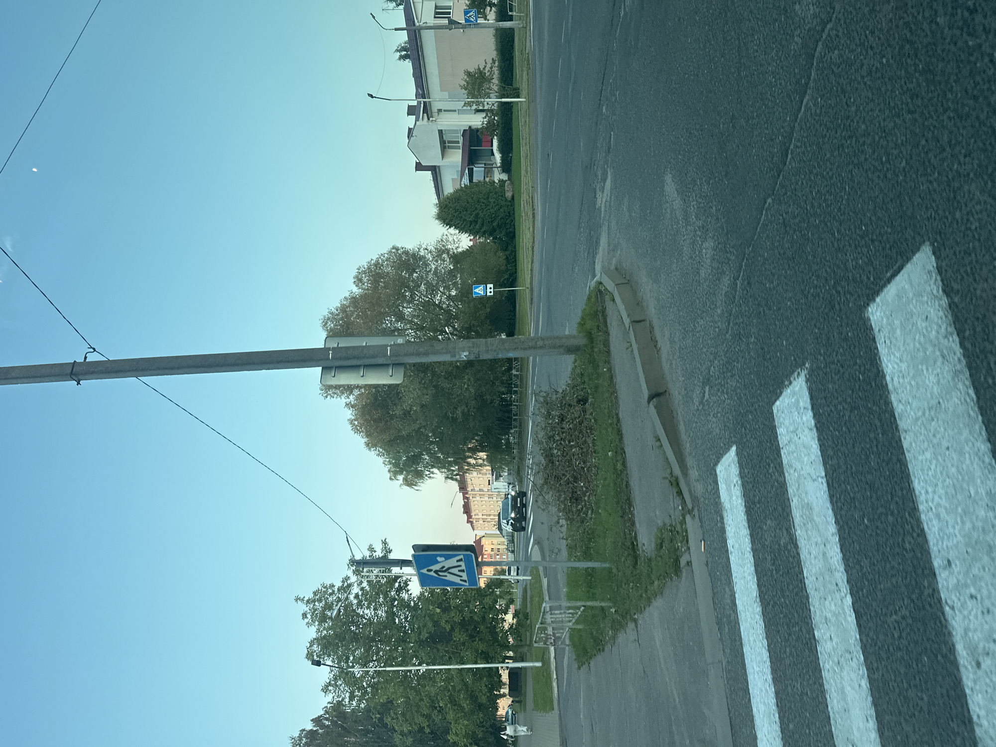 The image depicts a street scene in Maladzyechna, Belarus, on a clear day. In the foreground, a pedestrian crossing marked with white stripes is visible on asphalt. To the left, a sidewalk with a curb runs alongside the road. A metal barrier separates the sidewalk from a patch of grass and debris. A blue pedestrian crossing sign is mounted on a pole. Further down the street, several cars are present, including a black SUV driving towards the viewer and a white car driving away. Tall trees with green foliage line the street, and apartment buildings can be seen in the background. A lamppost is positioned near the pedestrian crossing sign. The sky is a clear, pale blue, with a faint crescent moon visible.