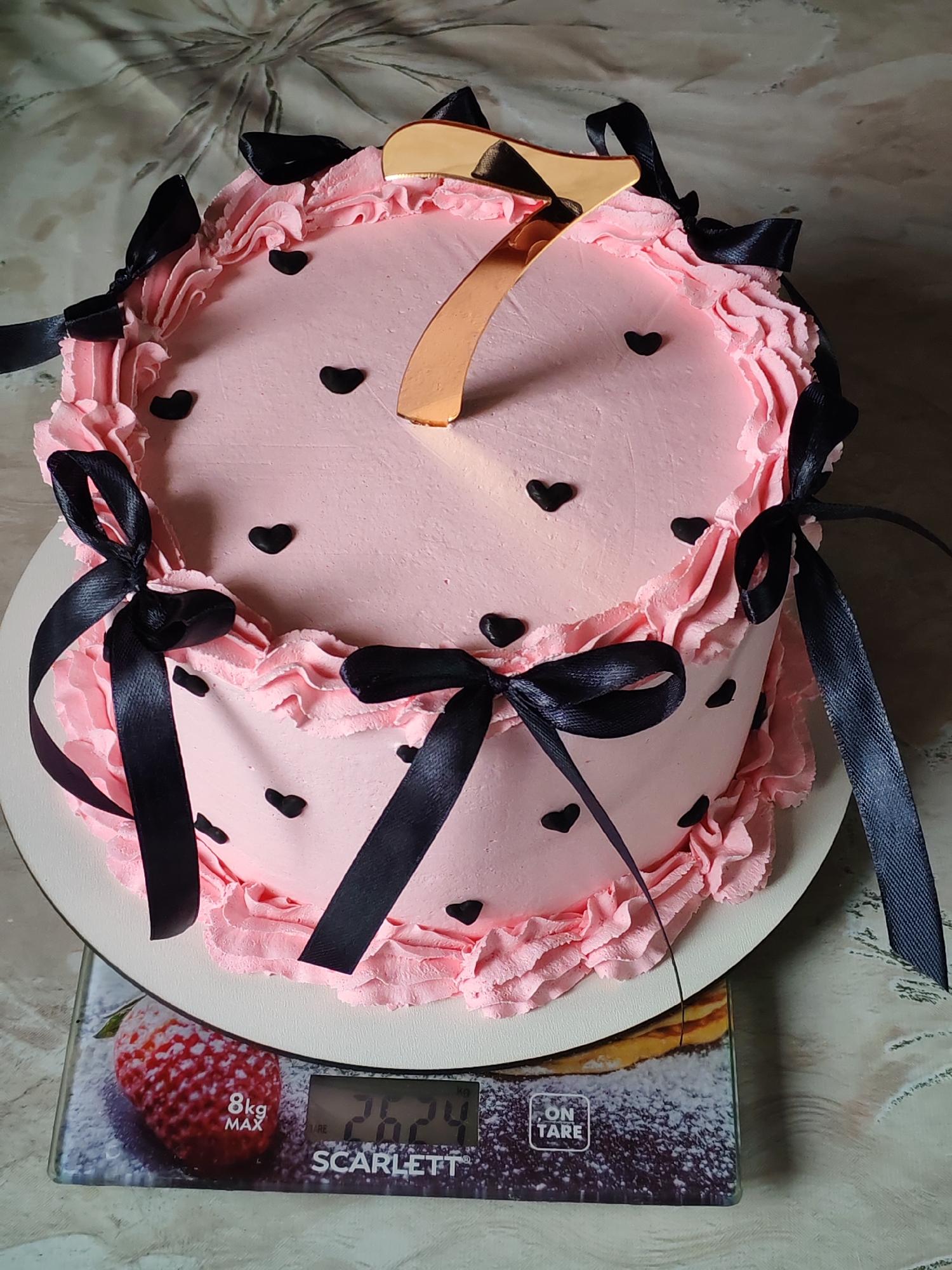 A pink cake with black heart decorations and black ribbon bows is placed on a white cake board, which in turn rests on a digital scale. The cake has a ruffled pink frosting border and a gold number "7" cake topper. The digital scale shows a weight of "2624" grams. The background is a patterned fabric. The scene is indoors.