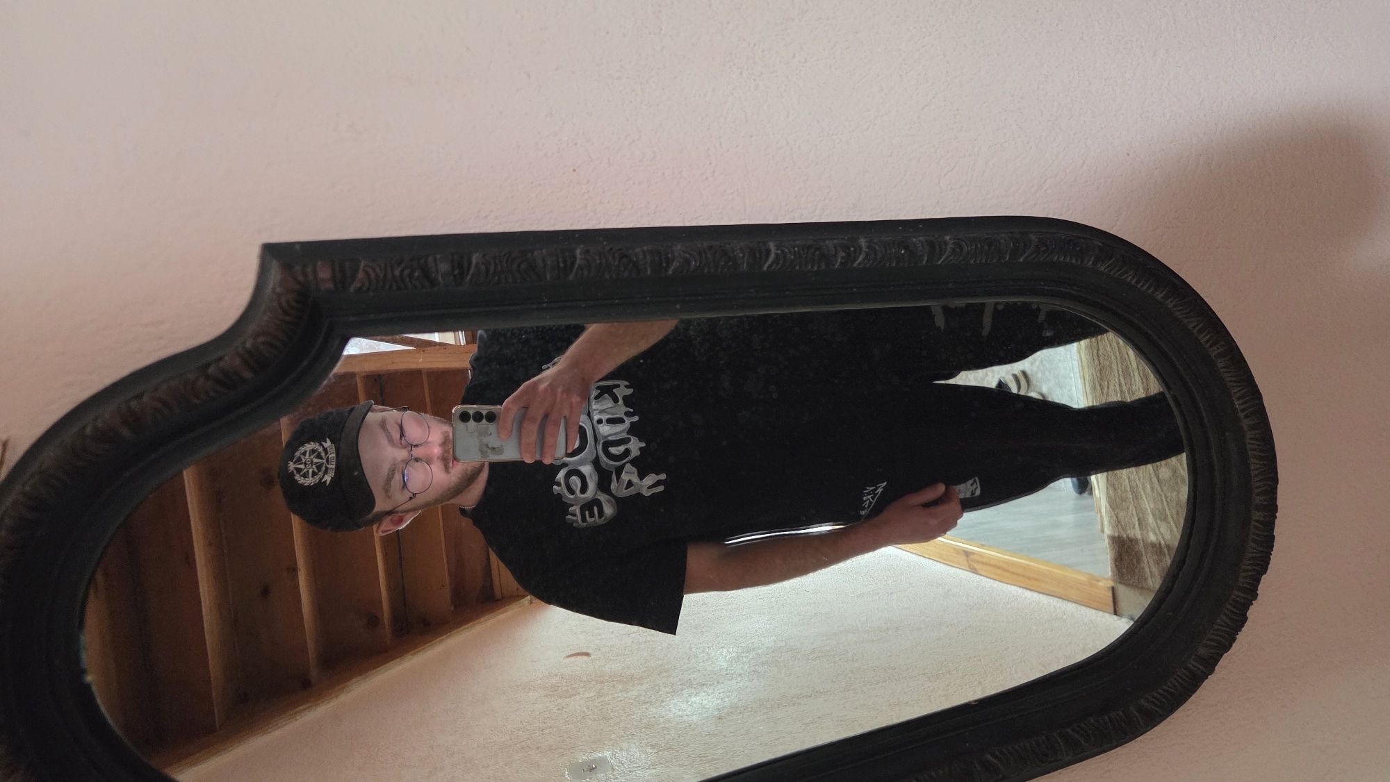A man wearing a black t-shirt, black pants, glasses, and a black hat stands in front of a mirror. He is holding a cell phone in his right hand and taking a selfie. The mirror is oval-shaped with a black ornate frame. The background shows a white wall, stairs, and a door. The location appears to be indoors. It is impossible to ascertain if this is the city of Bania, Bulgaria.