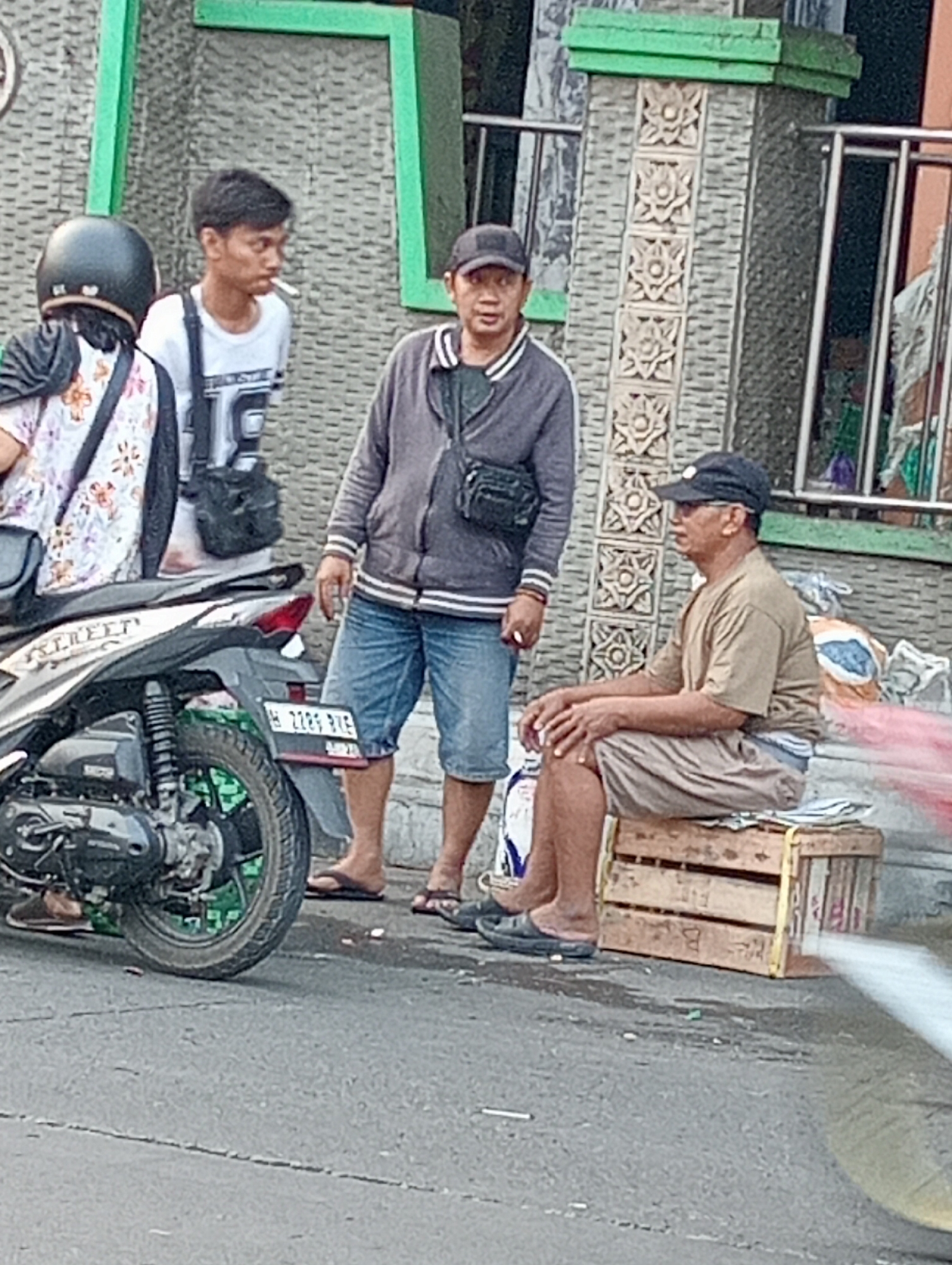 The image depicts an outdoor scene on a street in Demak, Indonesia. Three men are present. In the foreground, a man wearing a dark cap, glasses, and a light brown t-shirt and shorts sits on a wooden crate. To his left, another man in a gray jacket, blue denim shorts, and a black cap stands and appears to be smoking. To the far left, a person wearing a helmet, a patterned t-shirt, and a headscarf is partially visible, standing near a motorcycle. The motorcycle is parked on the asphalt road, which shows signs of wear and scattered debris. Behind the individuals, a building with patterned stone accents and green architectural features is visible. The overall setting suggests a casual, everyday street environment.