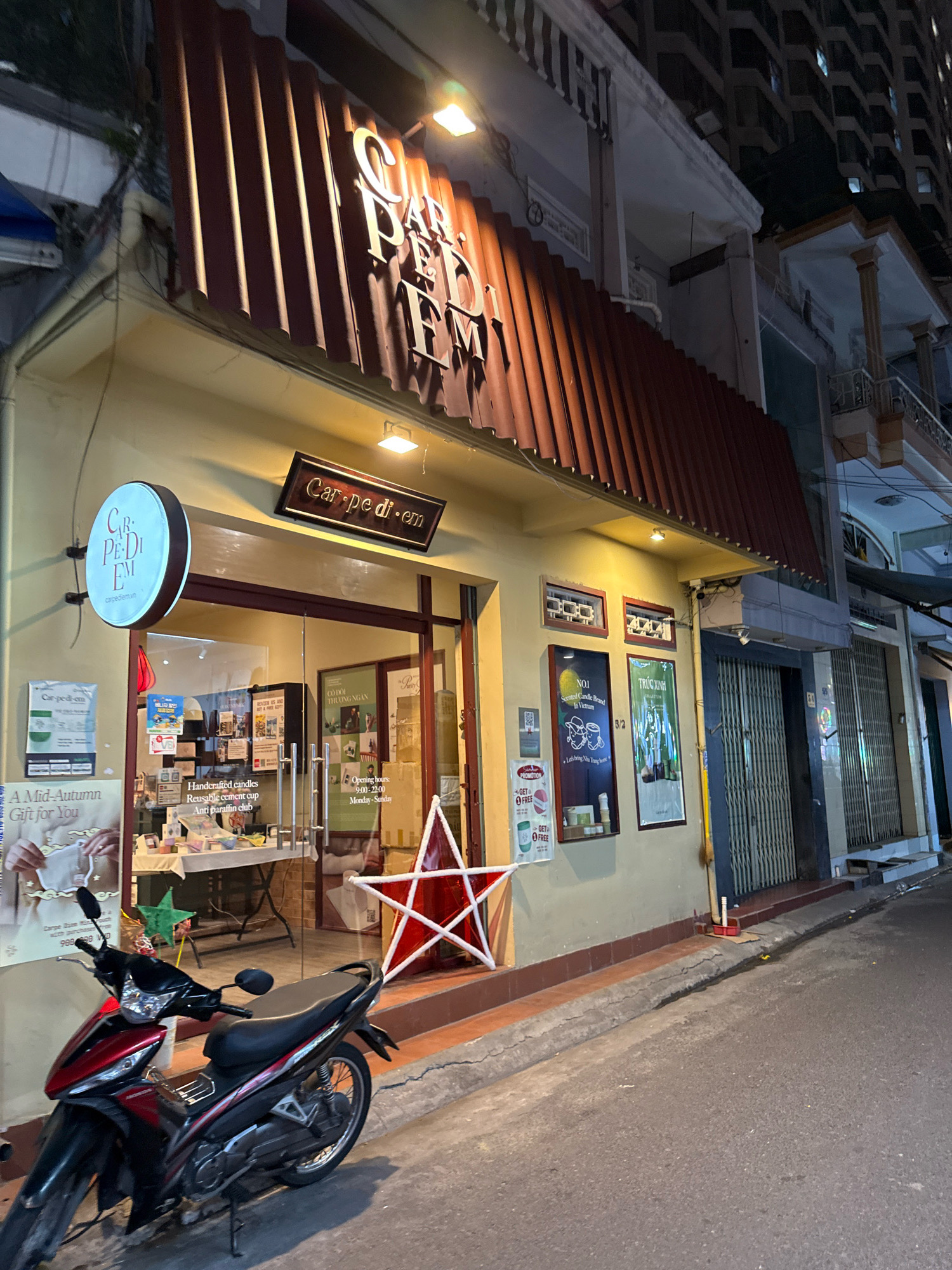 The image captures the exterior of a shop named "Carpe Diem" at night in Nha Trang, Vietnam. The shop features a prominent red corrugated metal awning with the store name "Carpe Diem" displayed in raised white letters. A circular illuminated sign with "Carpe Diem" is mounted to the left of the entrance, and a rectangular wooden sign with "Carpe Diem" is above the shop's entrance. The shop's facade is painted a light yellow, with large glass windows and doors revealing merchandise inside. Displays and posters are visible through the windows, including information about "Handcrafted candles," "Reusable cement cup," and "Anti paraffin club." A red and white star decoration is positioned outside the shop entrance. A red and black motorcycle is parked on the sidewalk to the left of the shop. The street in front of the shop is paved, and buildings with multiple stories are visible in the background, suggesting an urban environment. The lighting indicates it is nighttime, with artificial lights illuminating the shop's signage and interior.