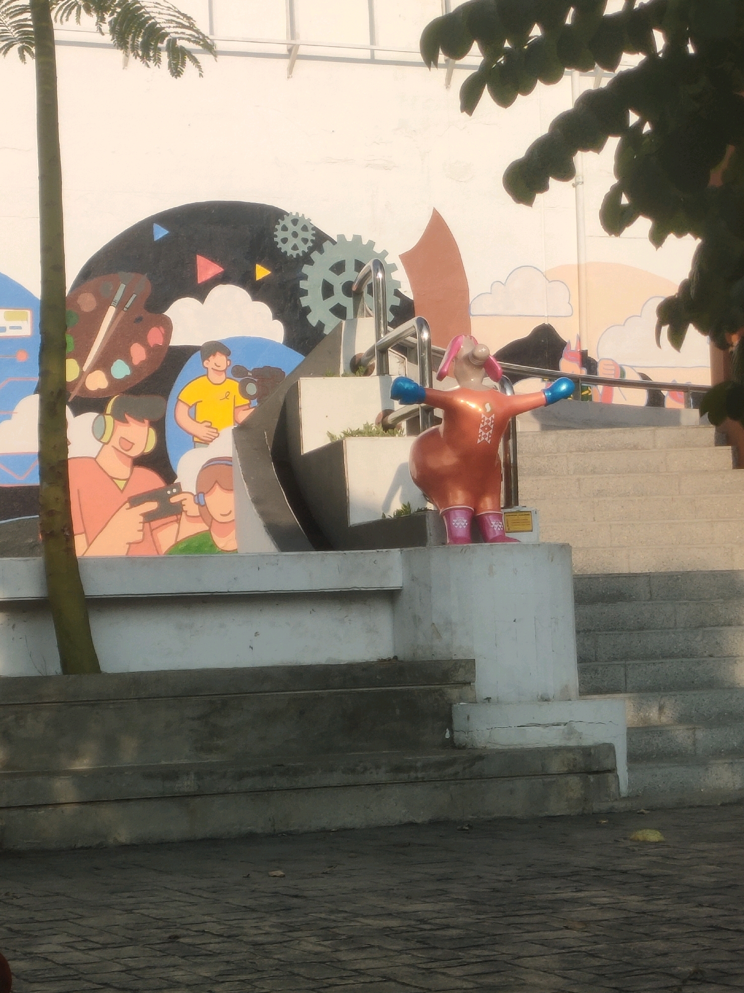 The image depicts an outdoor scene in Surabaya, Indonesia, featuring a mural on a white wall and a quirky statue. The mural showcases stylized figures engaged in activities like painting, gaming, and filming, with elements like a palette, headphones, a camera, and gears incorporated into the design. In front of the mural, a vibrant orange statue of a character with pink ears, blue gloves, and pink boots stands with its arms outstretched. The statue is positioned on a concrete pedestal. Behind the statue, a set of concrete stairs ascends, with a metal railing. The surrounding environment includes a tree trunk on the left, with some foliage obscuring the upper part of the image, and a paved ground area with cobblestone patterns in the foreground. The lighting suggests it is daytime, with sunlight casting shadows.