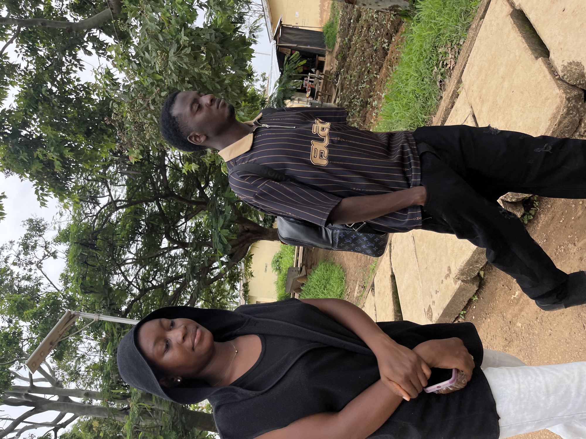 In the city of Jos, Nigeria, two young people, a woman and a man, walk side-by-side on a dirt path lined with concrete slabs. The woman, on the left, wears a black hooded vest over a black t-shirt and white pants. Her hands are clasped in front of her, holding a pink-cased mobile phone. The man, on the right, is dressed in a dark, pinstriped shirt with a tan collar and has his left hand in his pocket. He carries a backpack with a patterned strap. Behind them, a rural setting unfolds with lush green trees, a yellow building, and cultivated land. A solar-powered street lamp is visible above the woman. The overall atmosphere appears to be overcast.