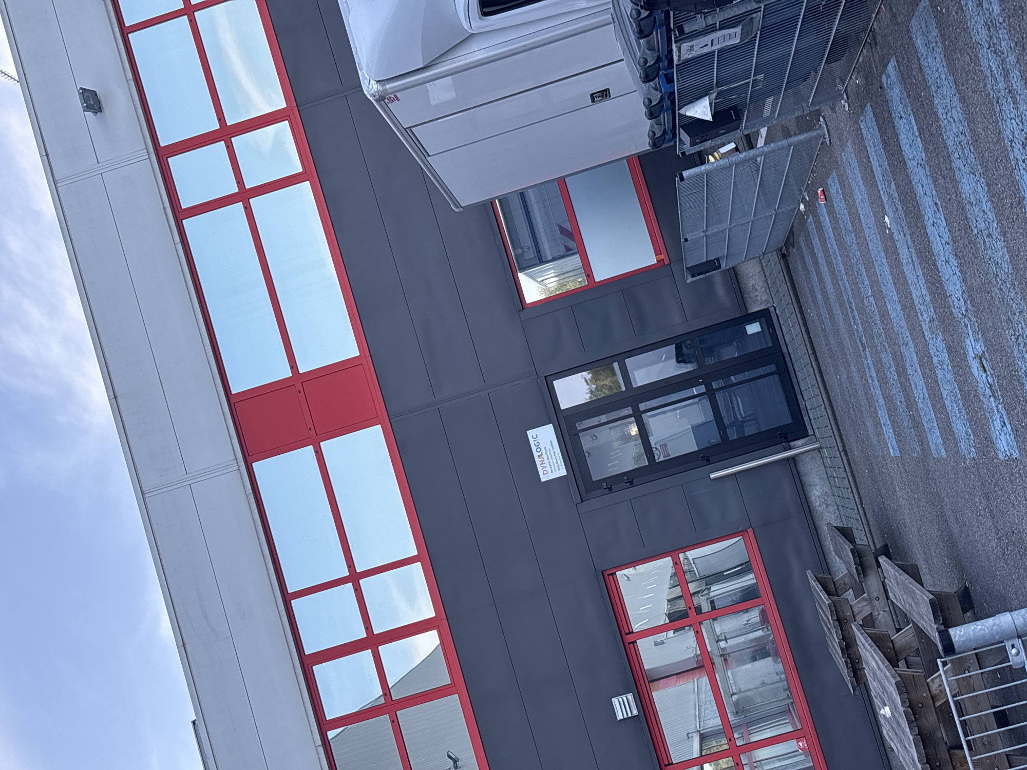 The image depicts the exterior of a modern industrial building with a dark gray facade and bright red window frames. The building has large, reflective windows that mirror the blue sky and some clouds. Above the dark gray section, there is a lighter gray upper level with a horizontal band. There is a glass entrance door on the dark gray section, with a small sign above it that reads "DYNALOGIC". A white truck with a cargo box is parked to the right of the entrance, partially obscuring some windows. In front of the building, there are wooden picnic tables and benches. The ground in front of the building is asphalt with blue painted stripes, possibly indicating a pedestrian crossing or parking area. Metal fencing is visible in the foreground and next to the truck. A single red and white can is on the ground near the fence. The scene appears to be in an industrial or commercial area, with a clear sky suggesting daytime. The image does not show any people. The specific location in the city of Boom, Belgium, is not directly indicated by any visible elements within the image itself.