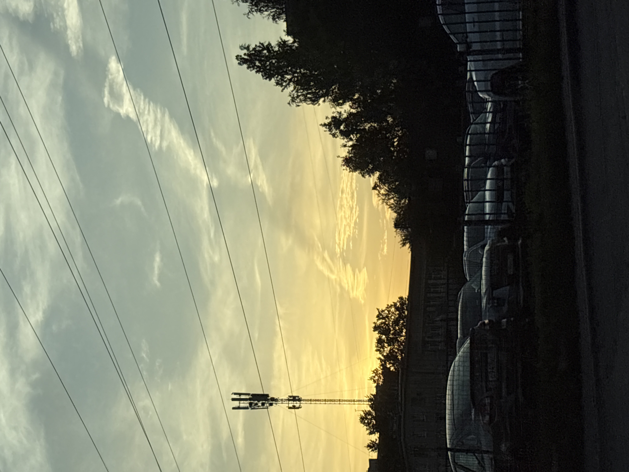 The image depicts a cell tower silhouetted against a dusky sky, with a row of parked cars in the foreground. The sky is a gradient of yellow and blue, with wispy clouds. Power lines stretch across the frame. The cell tower, located in Saint Petersburg, Russia, is a tall, metallic structure with multiple antennas. Behind the tower, trees and a building are visible. The cars, parked behind a metal fence, are mostly dark in color. The scene appears to be a typical urban landscape at dusk.
