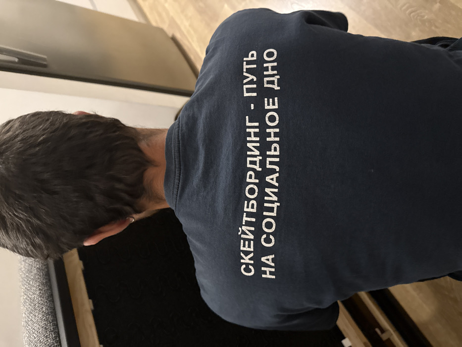 A man's back is visible, wearing a navy blue shirt. White text is printed vertically on the shirt, reading "СКЕЙТБОРДИНГ - ПУТЬ НА СОЦИАЛЬНОЕ ДНО" in Russian. The man has short, dark hair. The background includes a light-colored wooden floor and a gray object to the left, potentially a piece of furniture.