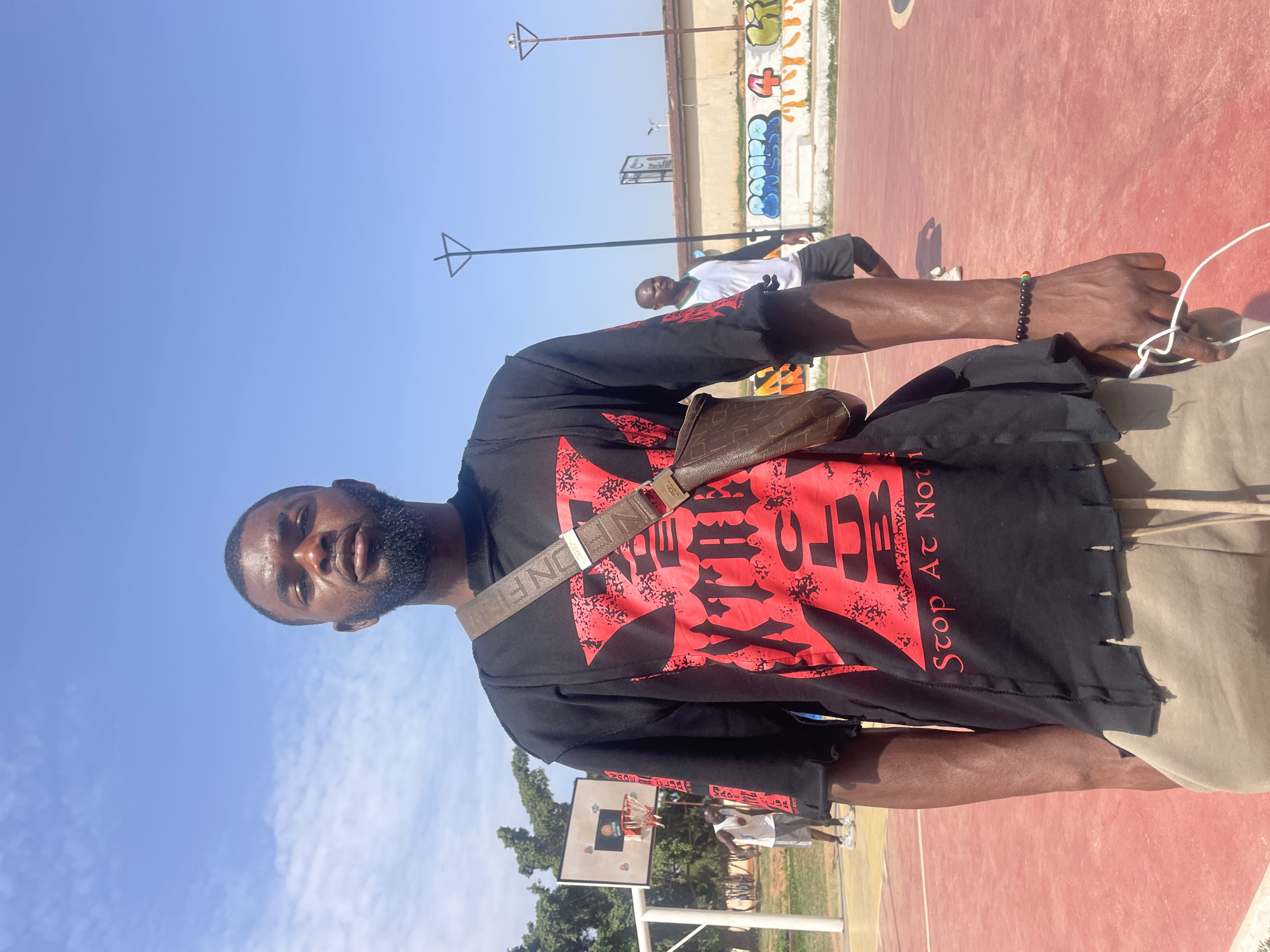 A man stands on an outdoor basketball court in Gura Top, Nigeria. The court is red, and a partial basketball hoop is visible in the background. A building with graffiti is in the distance. The man wears a black shirt with red graphics, khaki shorts, and a brown crossbody bag. Another man is visible in the background on the basketball court. The sky is blue with some clouds.